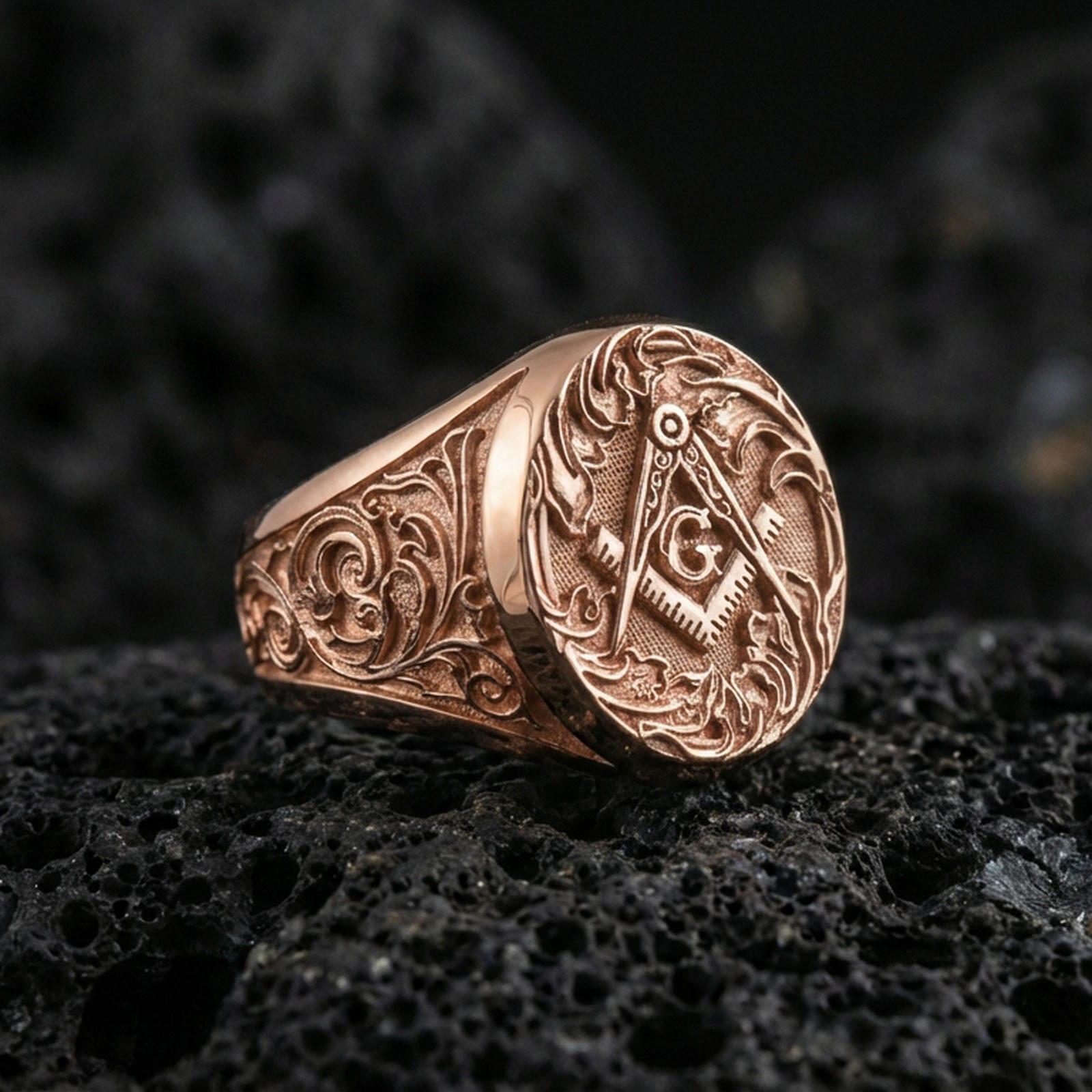 Rose gold plated Freemason square and compass ring with “G” symbol and detailed scroll engraving, macro photo on black volcanic rock