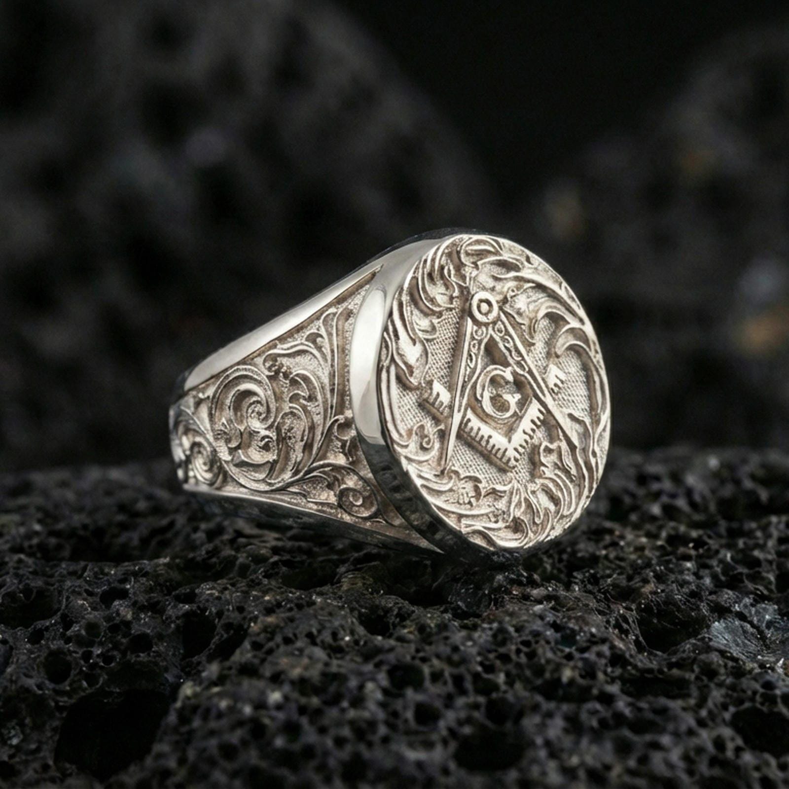 Polished silver Freemason ring with raised square and compass symbol and “G” letter, macro on volcanic stone background