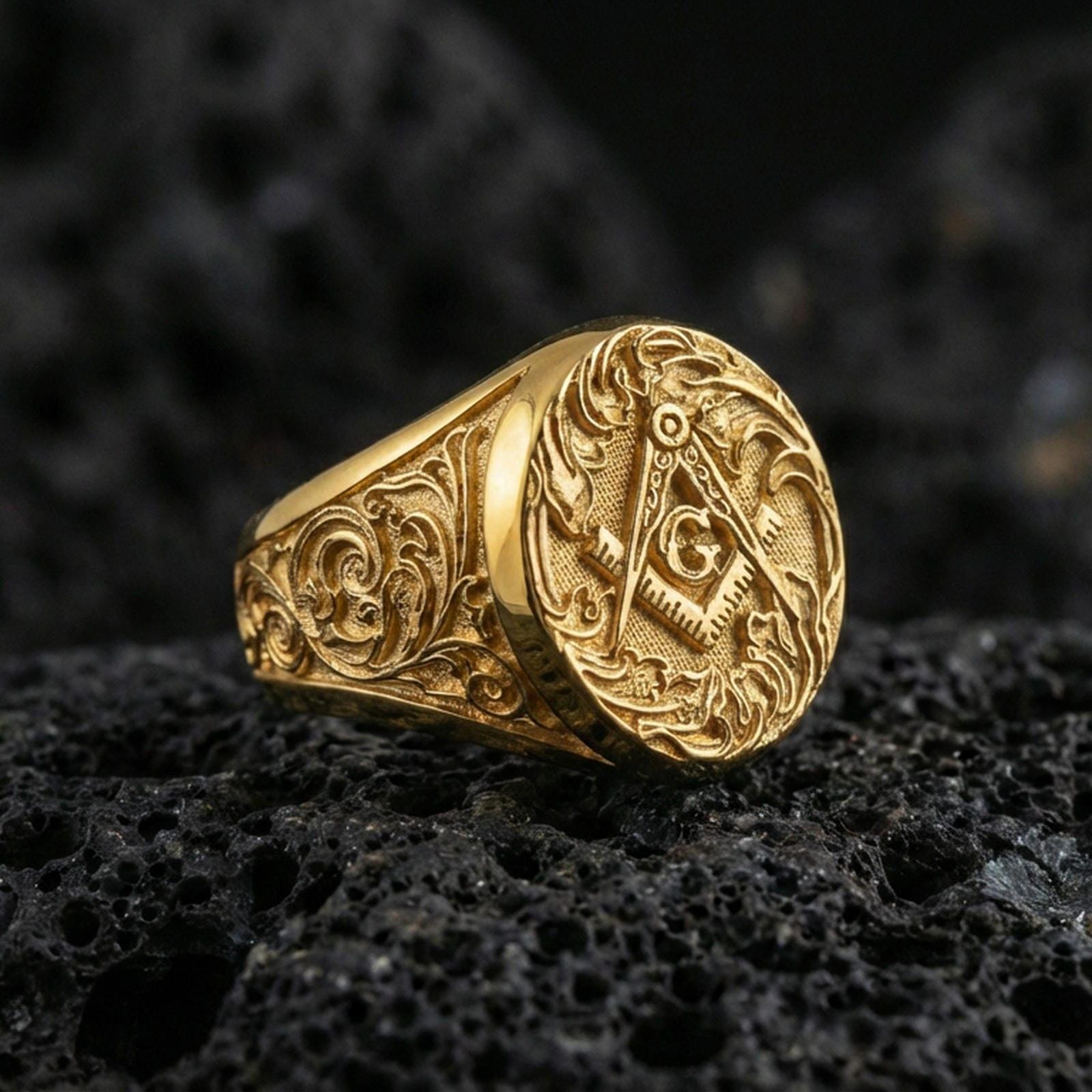 Gold plated Masonic signet ring featuring square and compass “G” emblem with engraved floral scroll band, close-up on volcanic rock