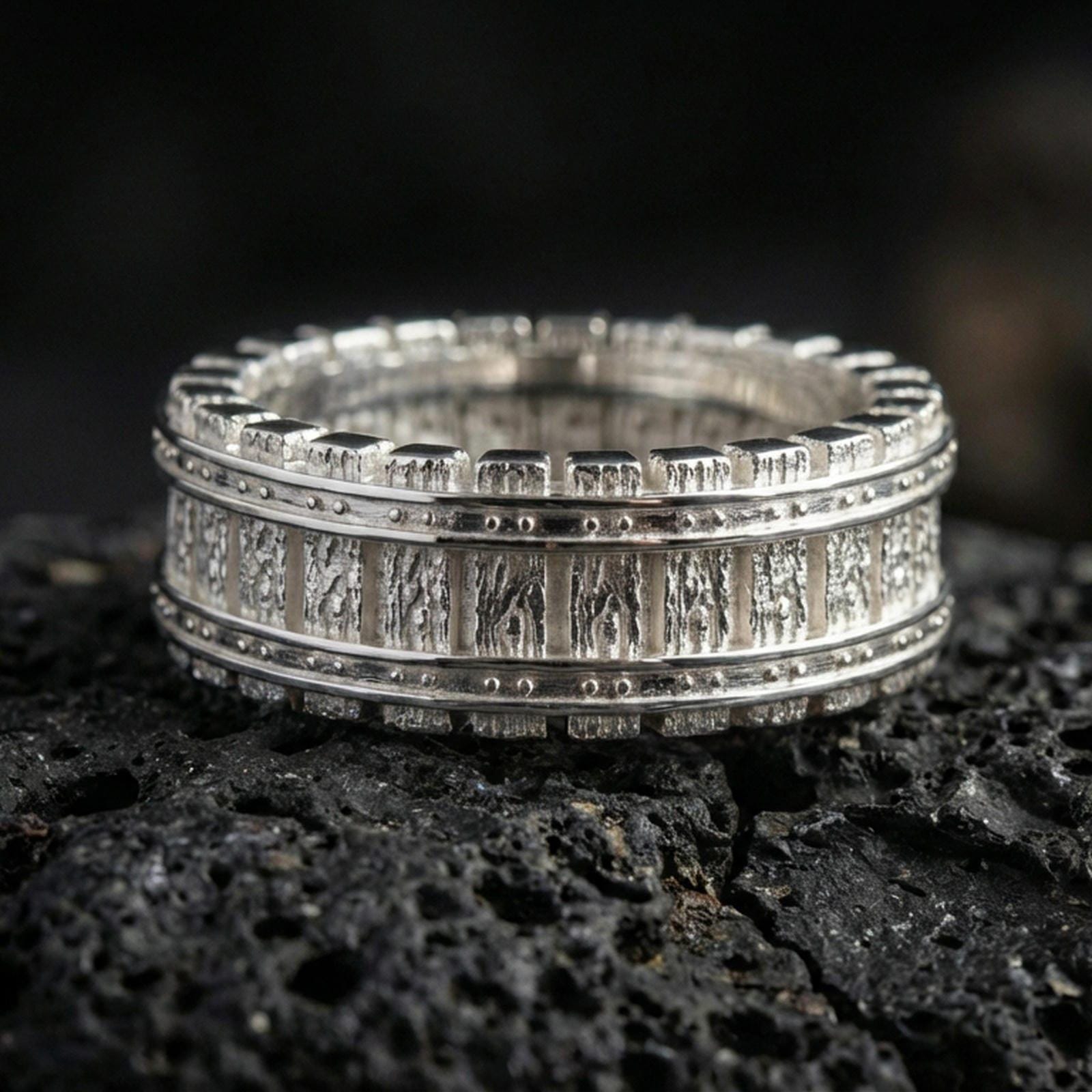 Polished silver industrial railway band ring on black volcanic stone, crisp rivet detailing and vertical track pattern, centered close-up studio lighting.