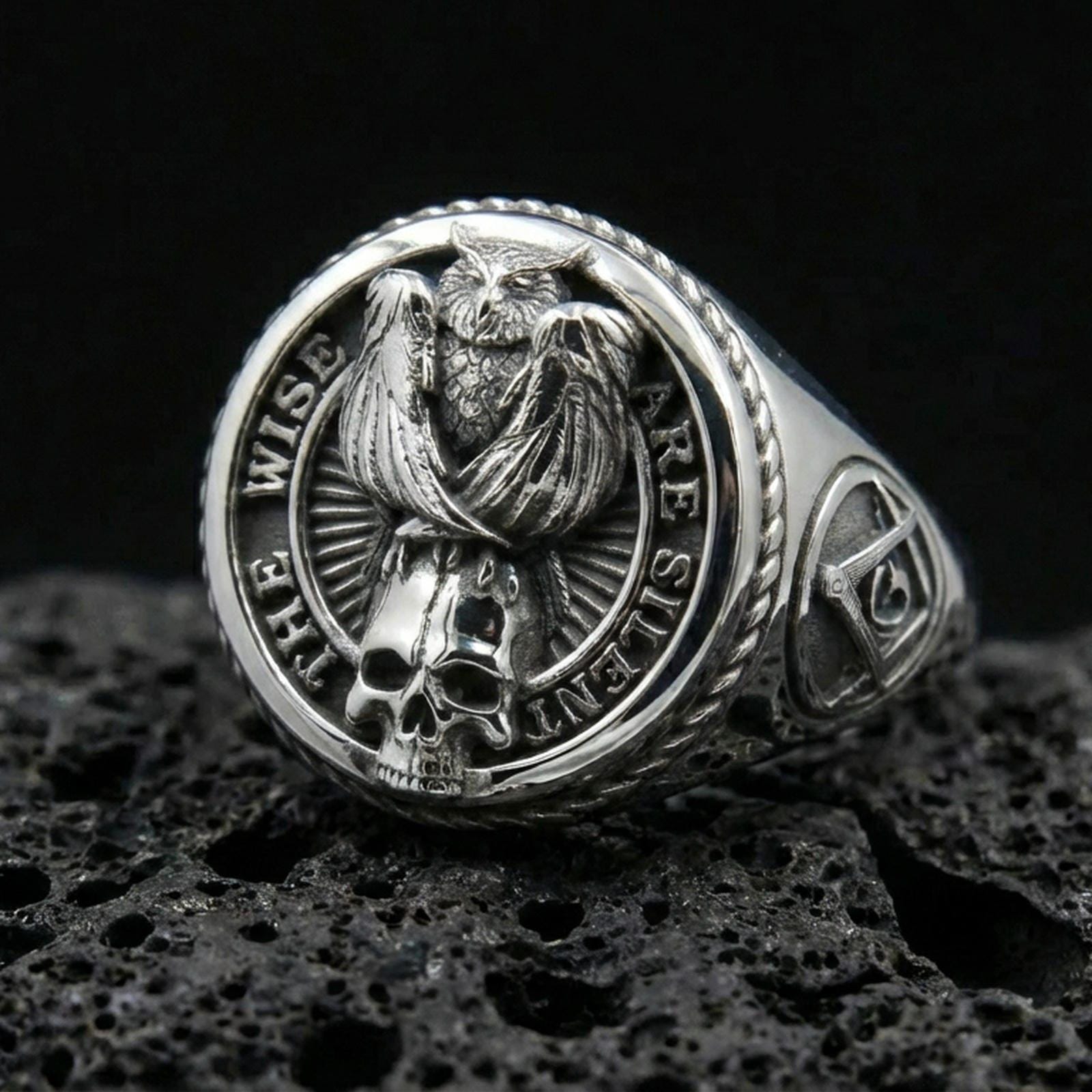 Polished silver signet ring featuring a detailed owl perched above a skull motif with engraved quote border, macro on volcanic stone