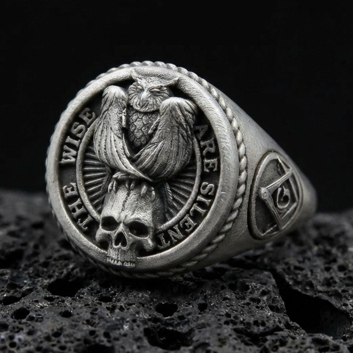 Oxidized sterling silver owl signet ring with skull detail and “THE WISE ARE SILENT” text, on black volcanic rock