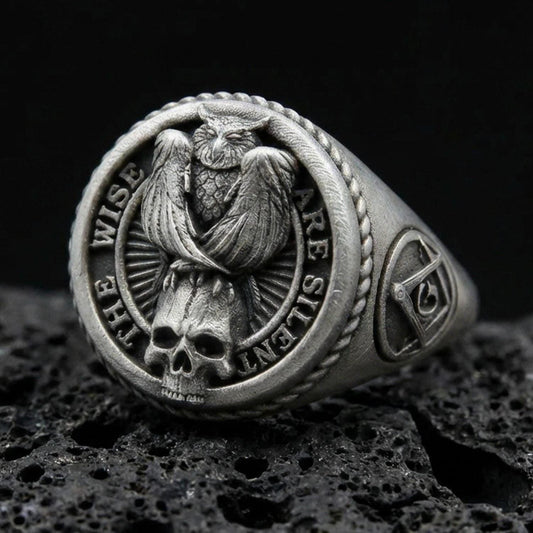 Oxidized sterling silver owl signet ring with skull detail and “THE WISE ARE SILENT” text, on black volcanic rock