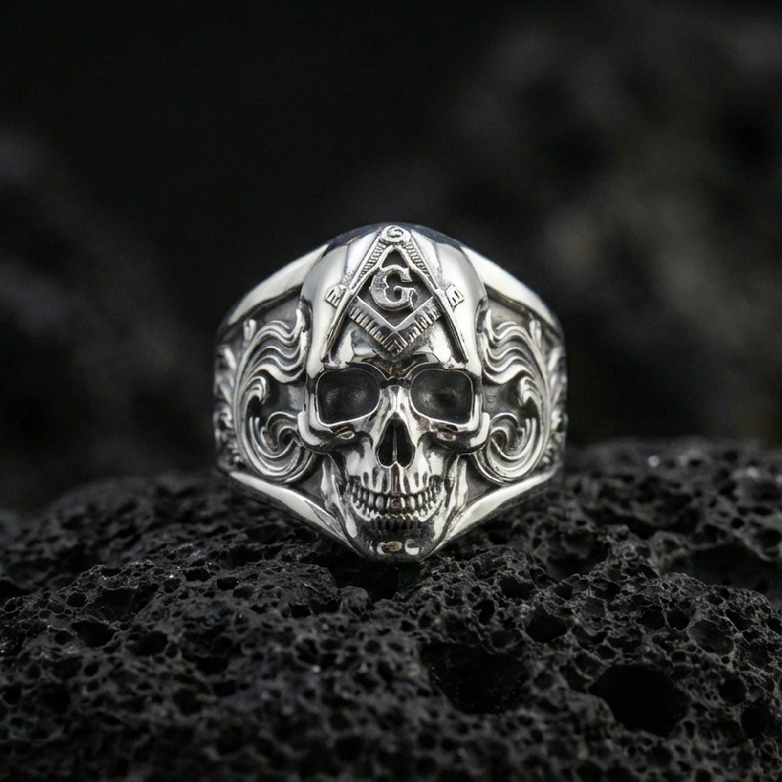 Polished silver Freemason skull signet ring featuring compass and letter G on the forehead with ornate side engravings, macro on volcanic stone