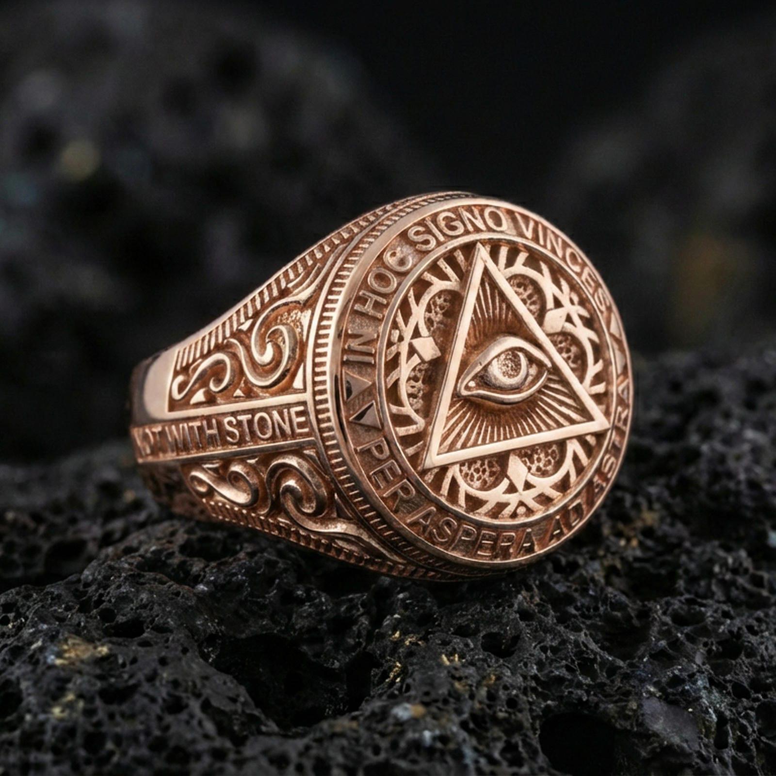 Rose gold plated Masonic eye triangle ring with engraved filigree sides and Latin inscription border, macro on black volcanic rock