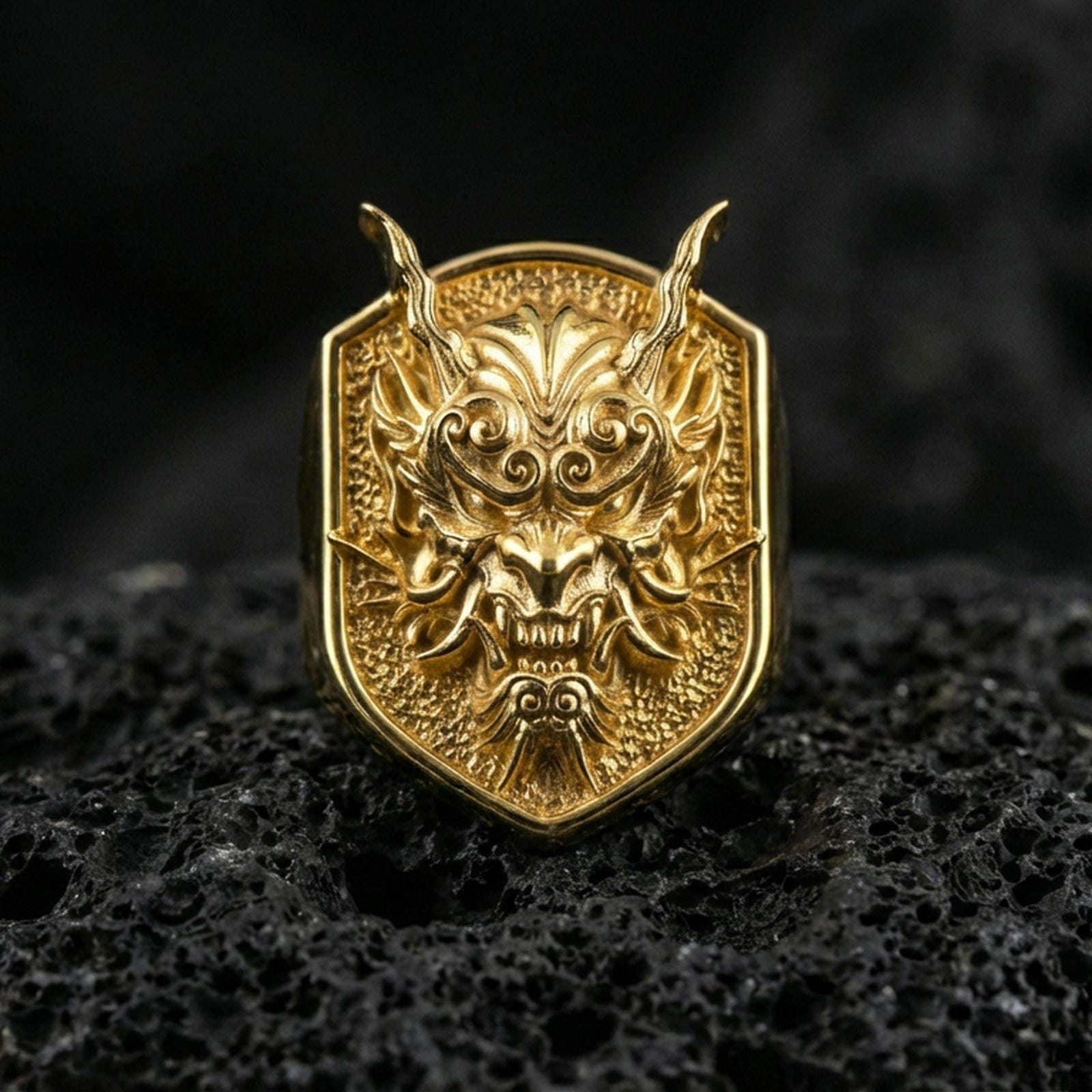 Gold plated oni mask signet ring featuring horned demon face carving and textured shield-shaped face, macro on volcanic rock