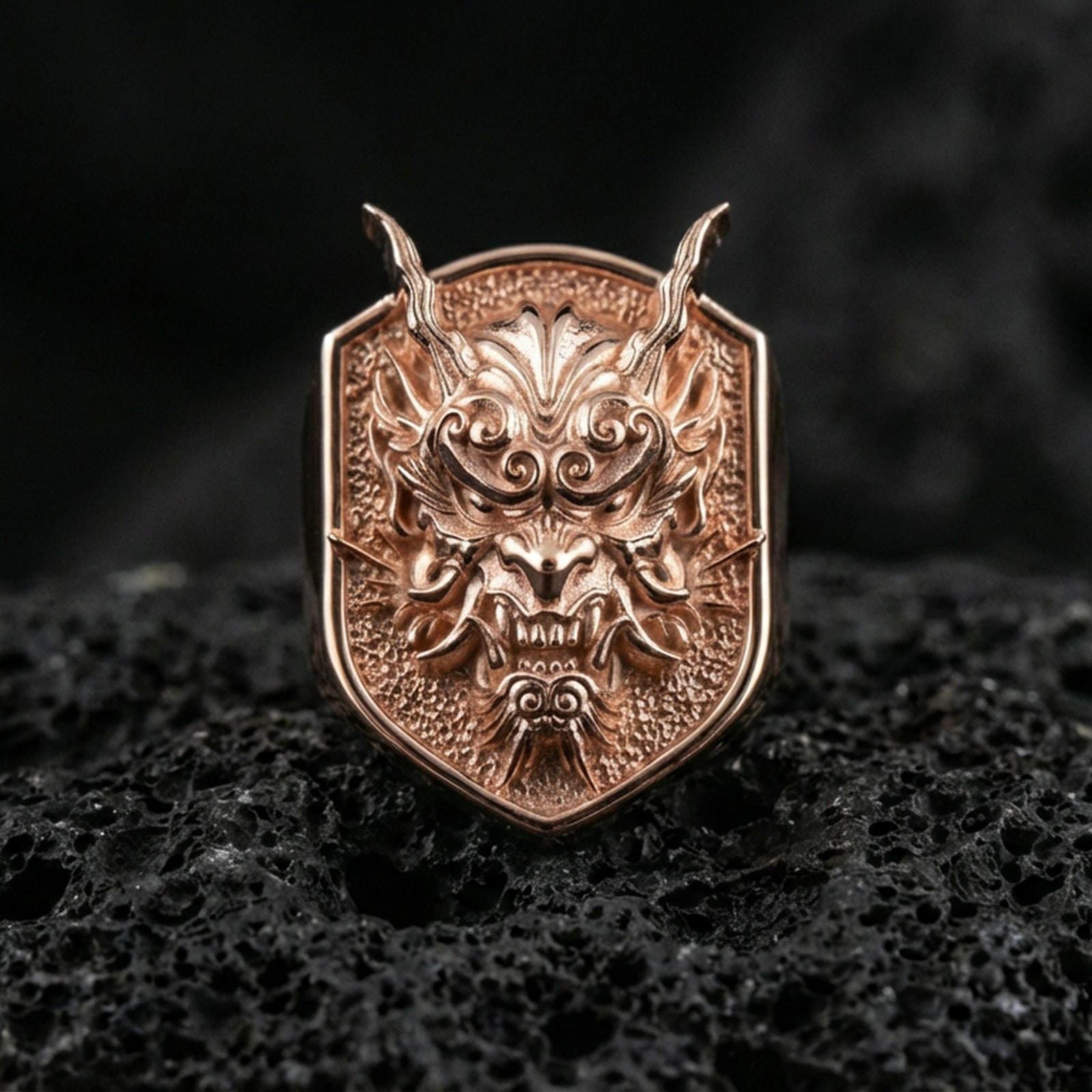 Rose gold plated Japanese oni mask ring with sculpted horns and fierce facial expression, close-up on black volcanic stone