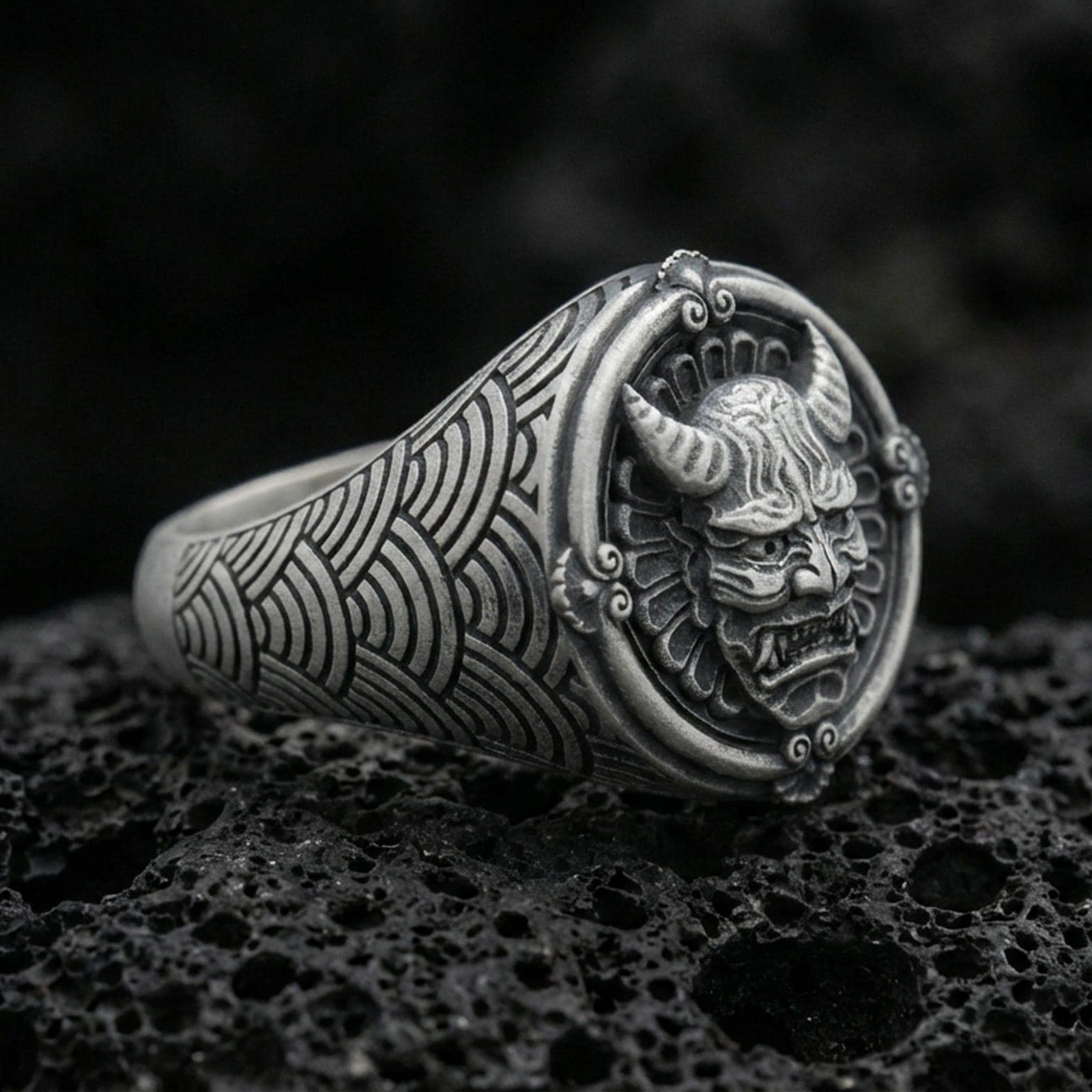 Oxidized sterling silver oni mask signet ring with horned Japanese demon face and seigaiha wave band engraving, macro on black volcanic rock