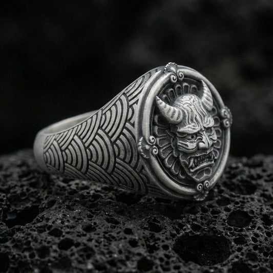 Oxidized sterling silver oni mask signet ring with horned Japanese demon face and seigaiha wave band engraving, macro on black volcanic rock