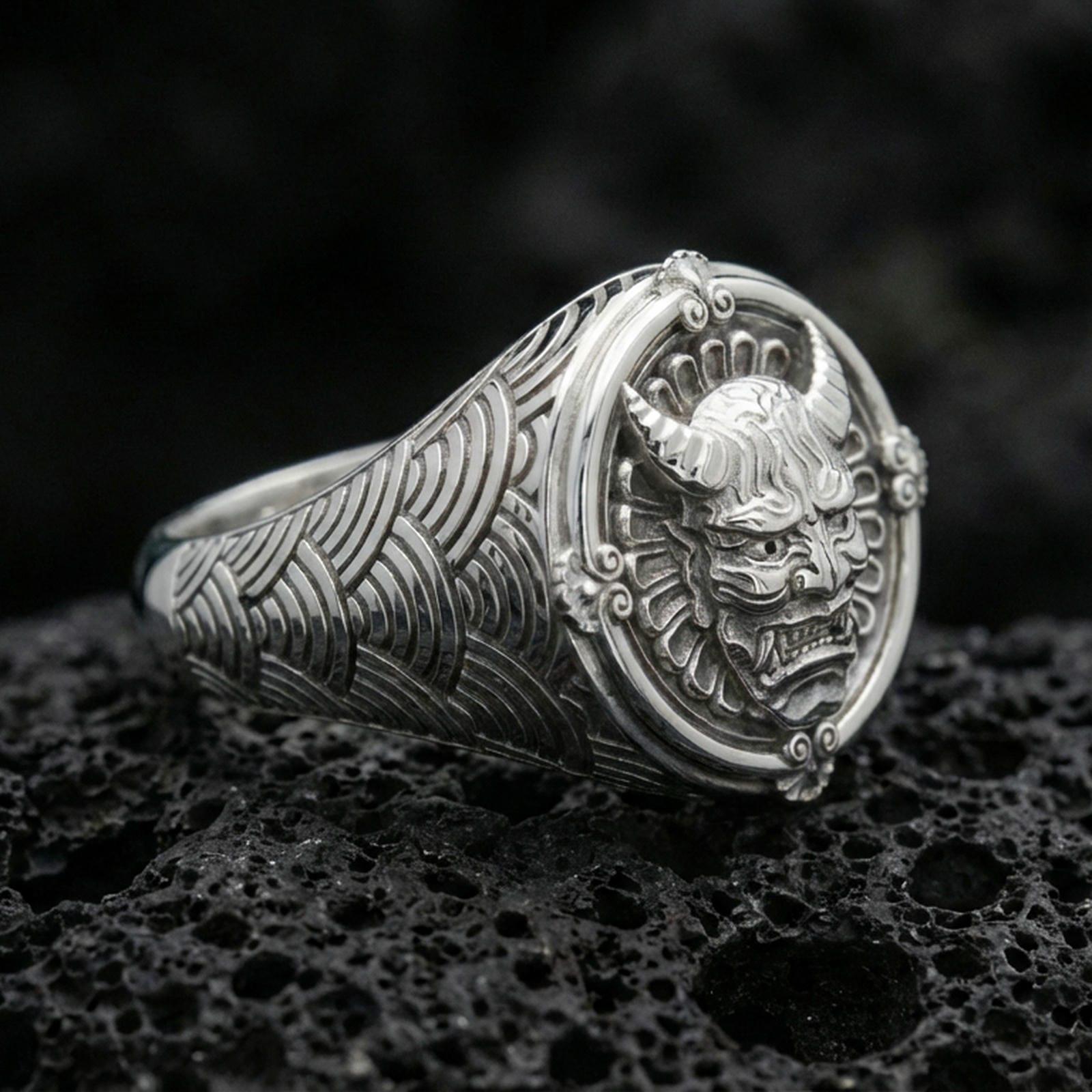Polished silver oni mask ring featuring seigaiha wave pattern shoulders and framed horned demon face, close-up on dark volcanic stone