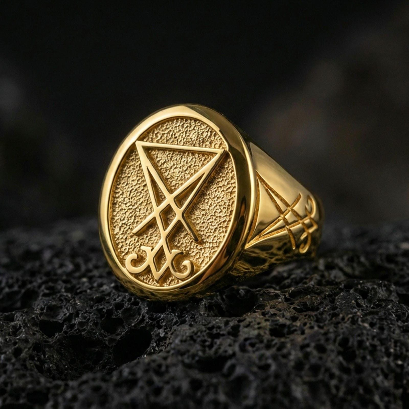 Gold plated Lucifer sigil signet ring with prominent raised occult symbol and dark textured background, macro on black volcanic rock