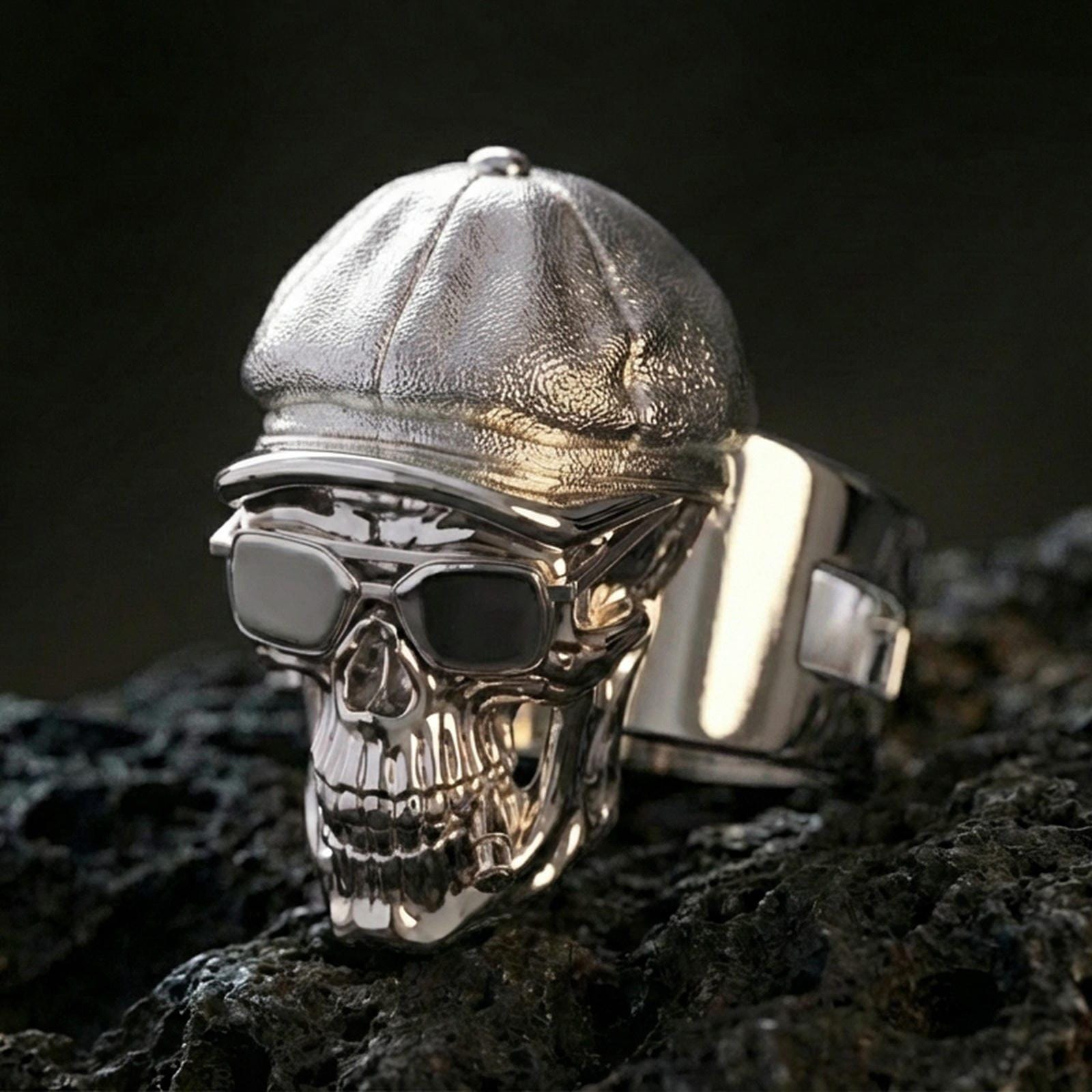 Sterling silver skull ring in a polished silver finish, featuring a cap, glasses, and detailed teeth relief, close-up on volcanic stone