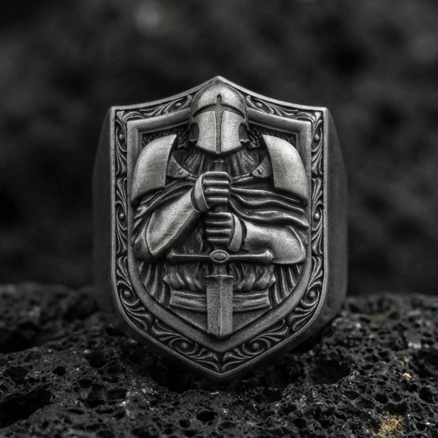 Sterling silver knight signet ring with an oxidized finish, shield-shaped face showing an armored warrior holding a sword, macro on black volcanic rock