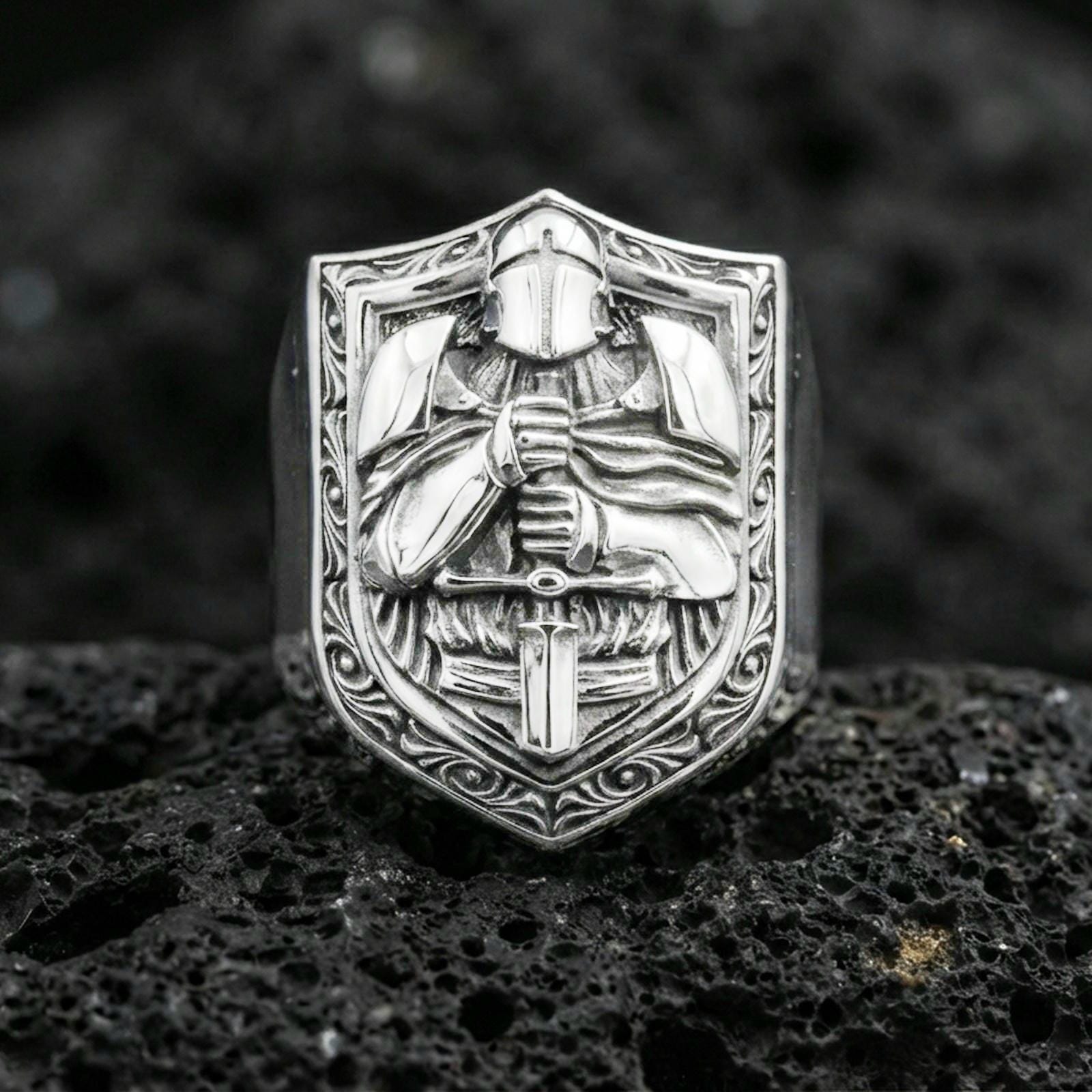Sterling silver knight signet ring in a polished silver finish, raised medieval warrior and sword relief on a shield face, close-up on volcanic stone