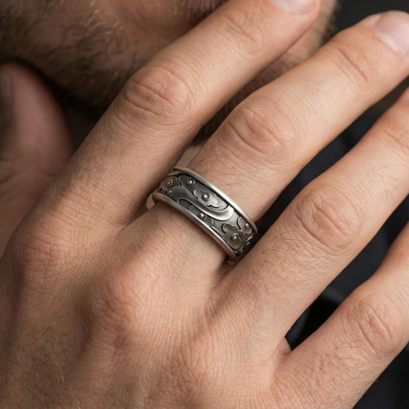 Oxidized sterling silver spinning cloud band worn on finger, whimsical swirl and flower relief detail on rotating center, natural lifestyle hand shot with shallow depth of field.