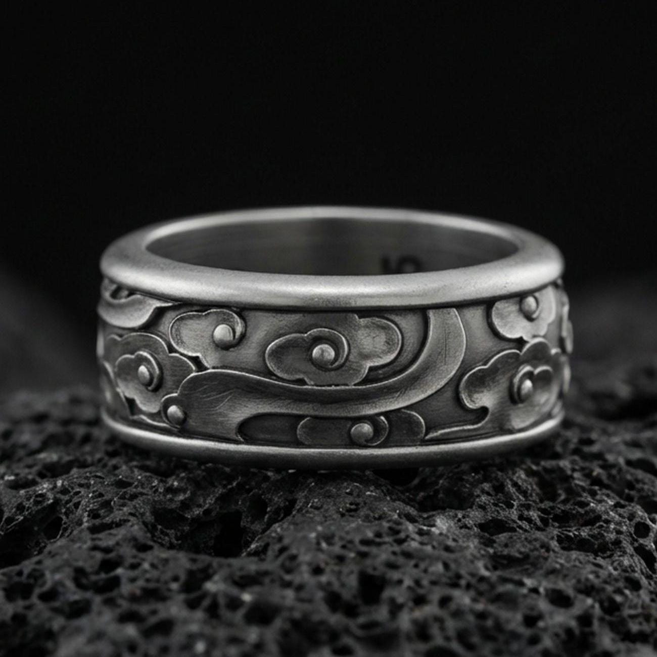 Oxidized sterling silver spinning cloud band ring on dark volcanic rock, whimsical swirl and flower relief on rotating center, premium macro product shot.