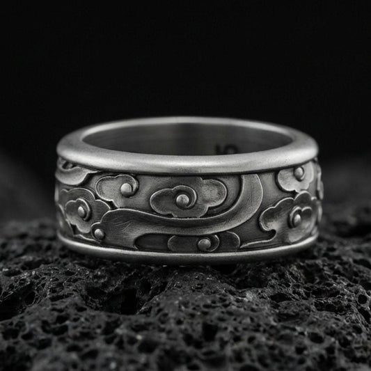 Oxidized sterling silver spinning cloud band ring on dark volcanic rock, whimsical swirl and flower relief on rotating center, premium macro product shot.
