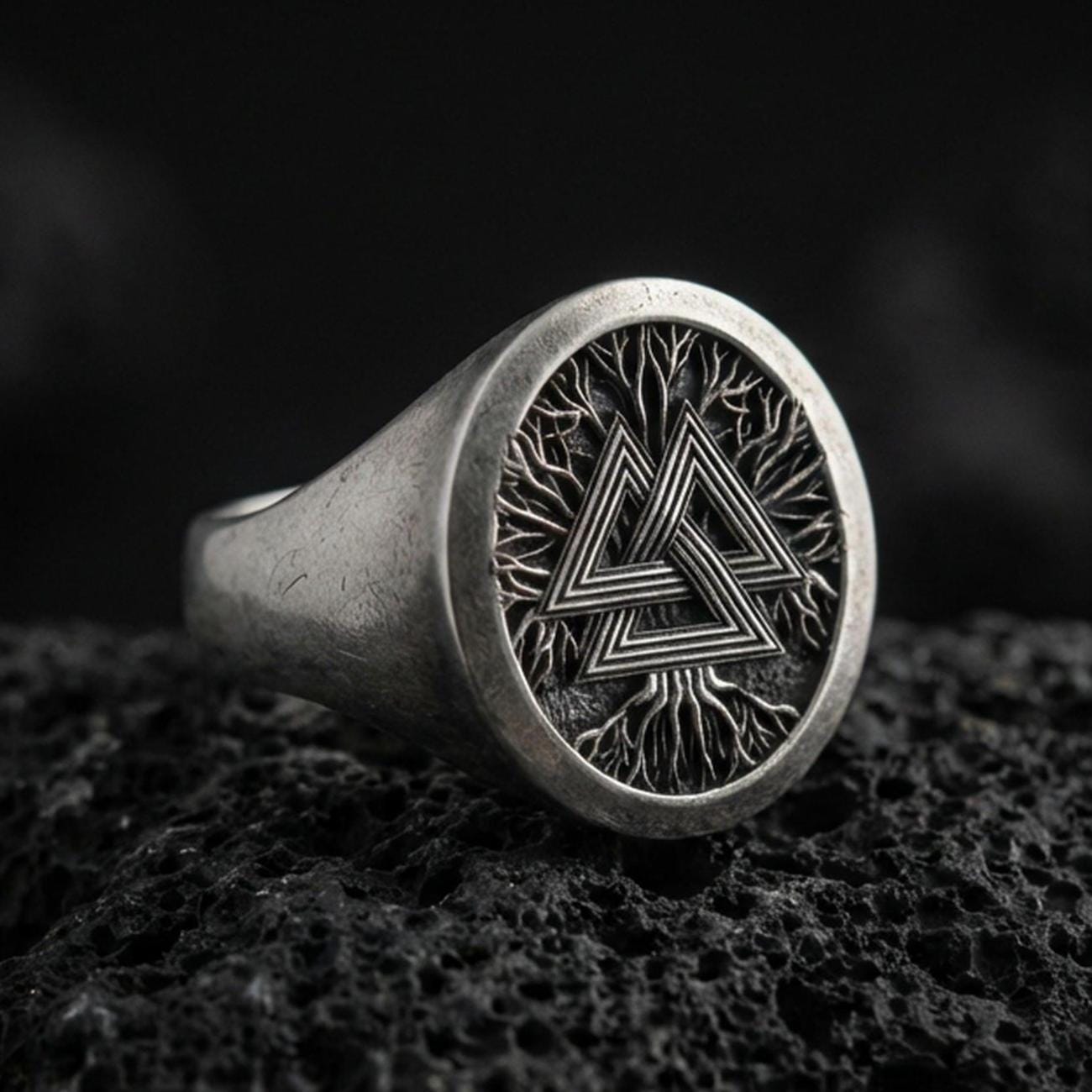 Sterling silver Valknut signet ring with an oxidized finish, round face featuring a Norse triple-triangle symbol over a branch-like texture, macro on black volcanic rock