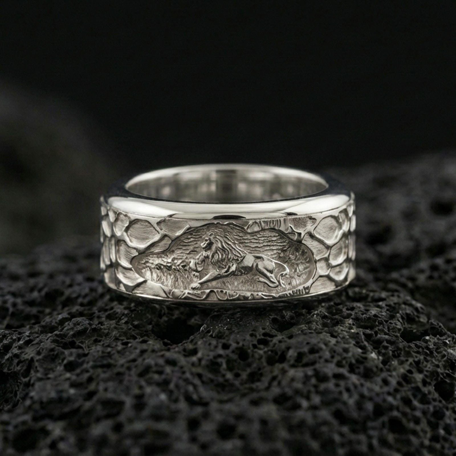 Polished silver lion scene spinner ring on black volcanic stone, crisp lion relief on rotating band with hammered-style texture, centered close-up studio lighting.