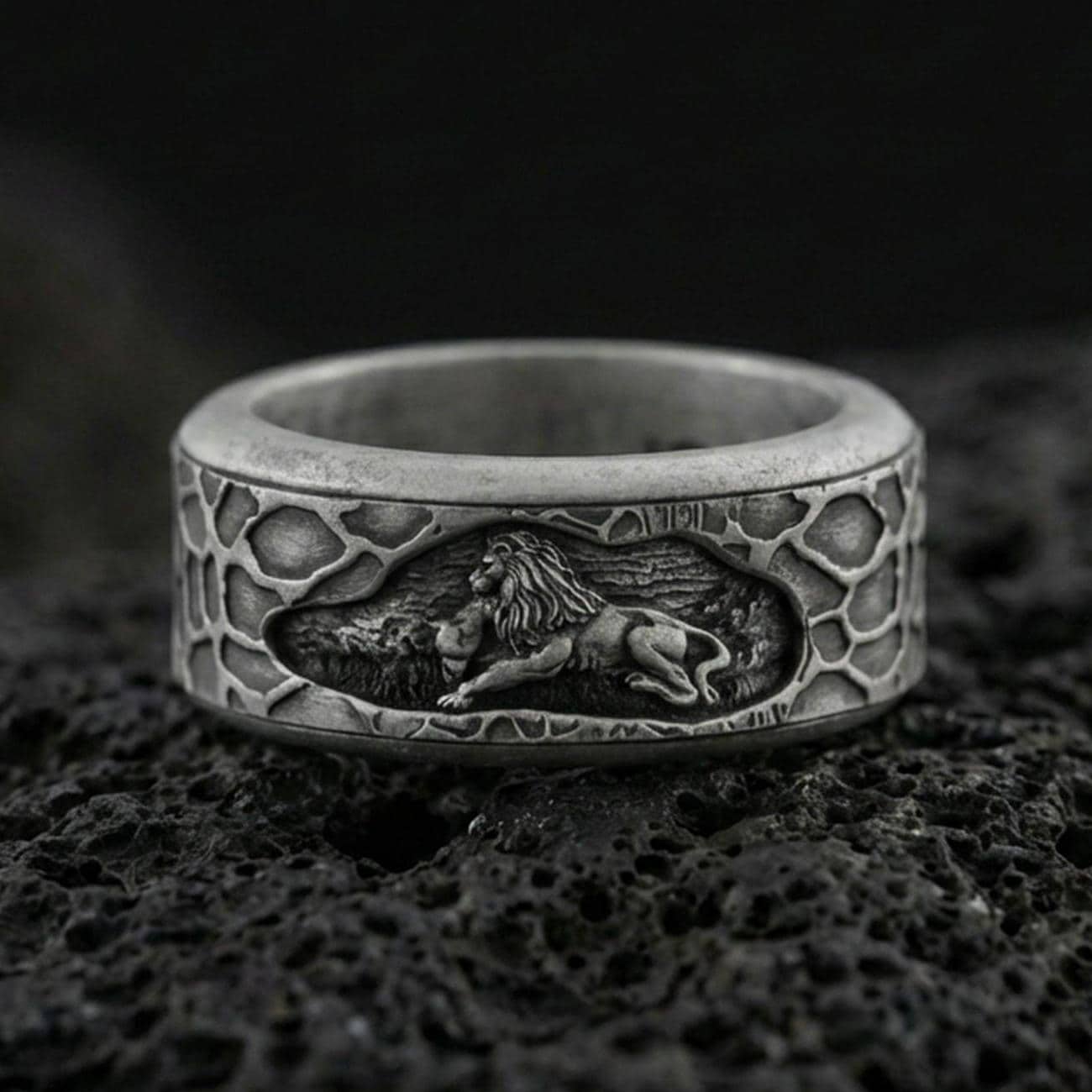 Oxidized sterling silver spinning lion band ring on dark volcanic rock, engraved lion scene on rotating center with textured border, premium macro product shot.