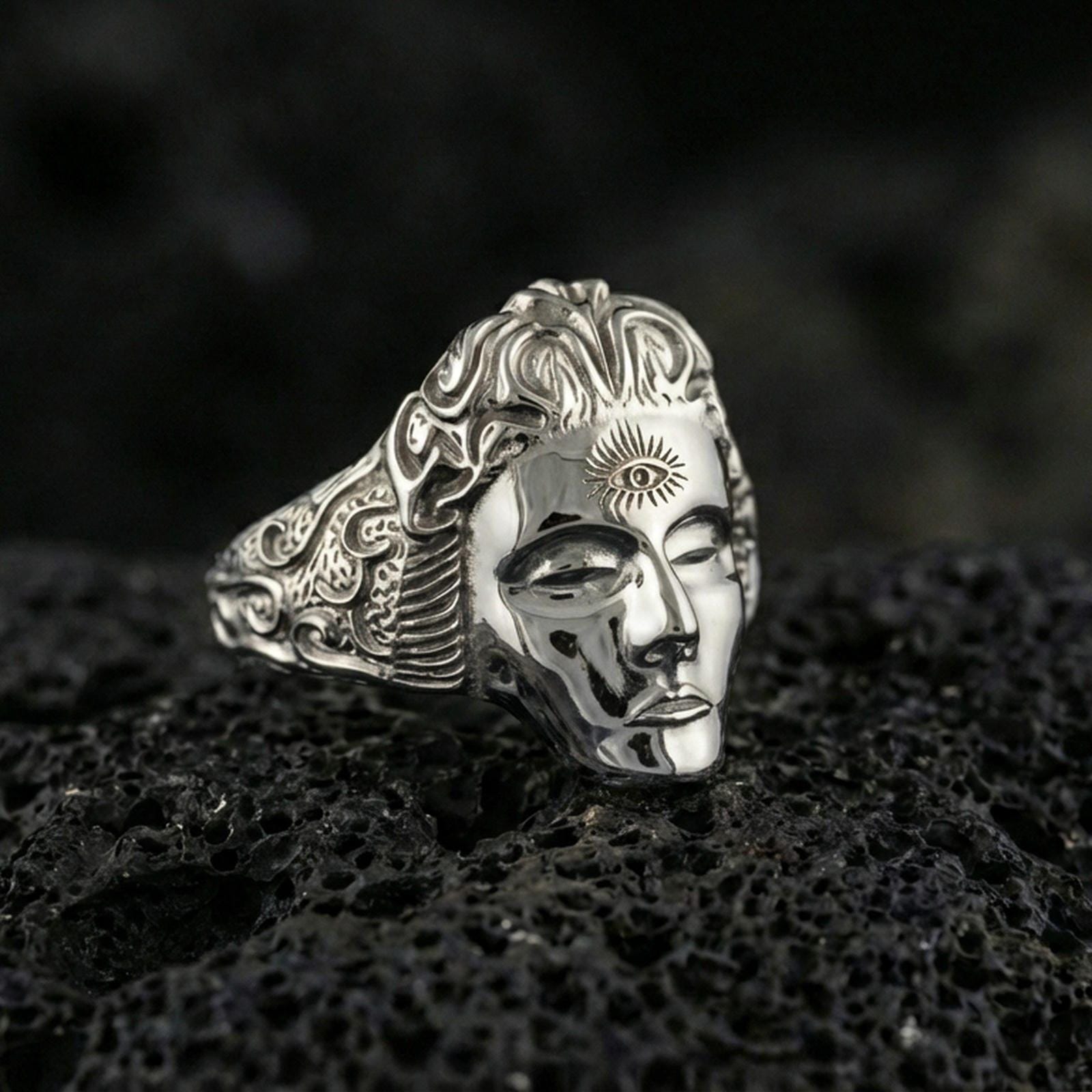 Sterling silver third eye face ring in a polished silver finish, raised mask profile with forehead eye symbol and ornate side engravings, close-up on volcanic stone