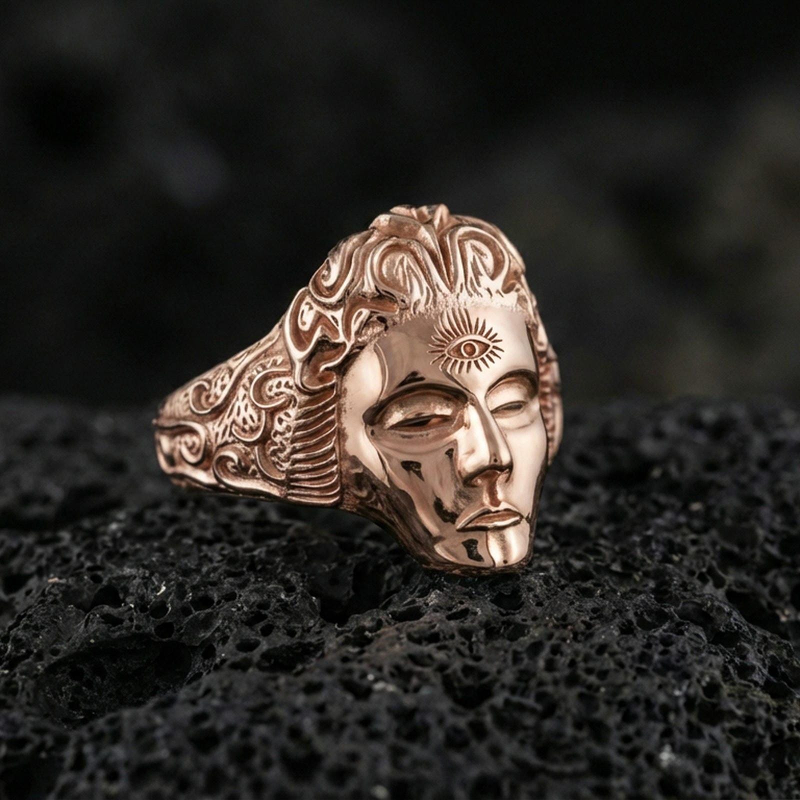 Sterling silver third eye face ring with a rose gold plated finish, sculpted face and third eye symbol with decorative engraved sides, close-up on volcanic rock