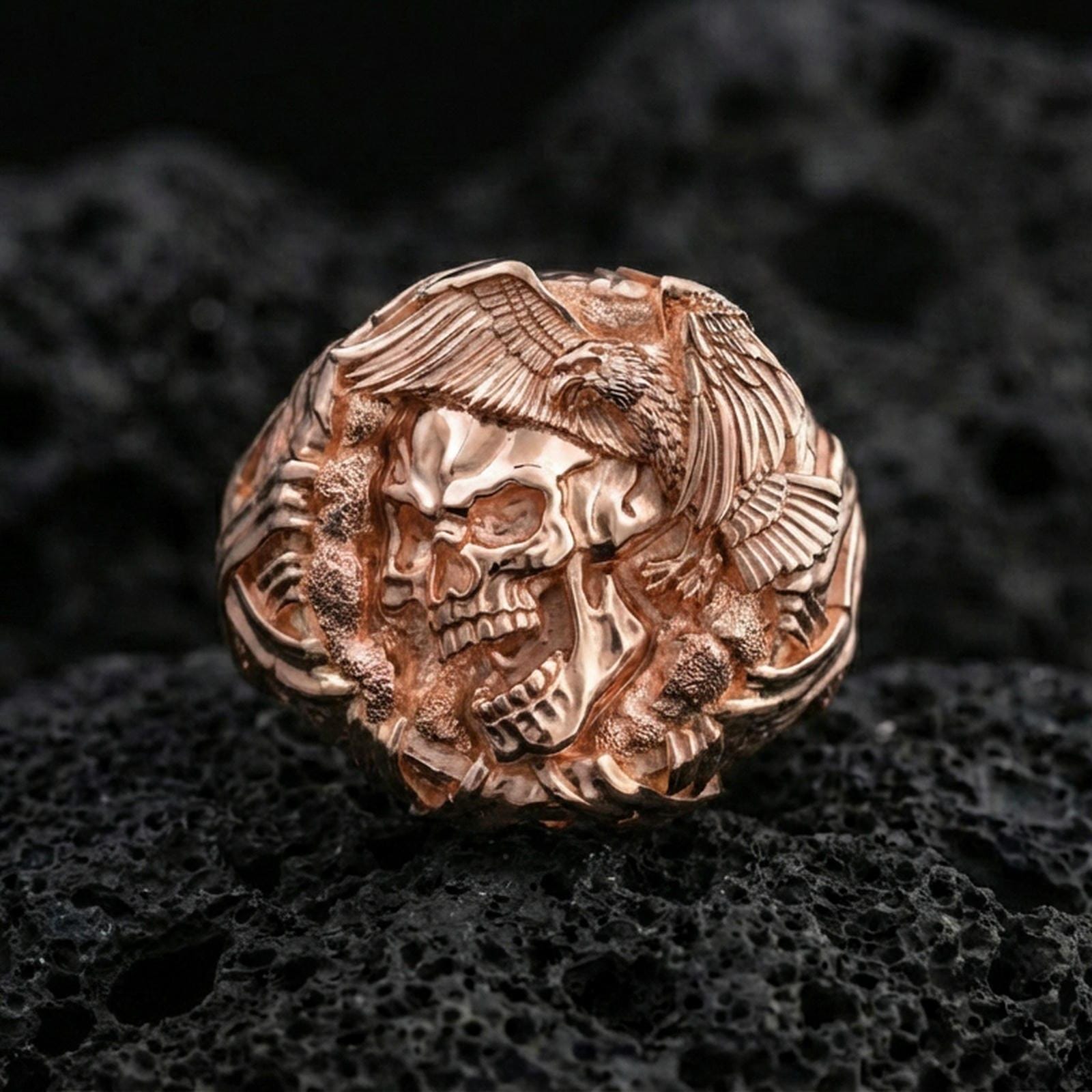 Sterling silver skull ring with a rose gold plated finish, detailed skull and eagle motif with sculpted wings, close-up on volcanic rock