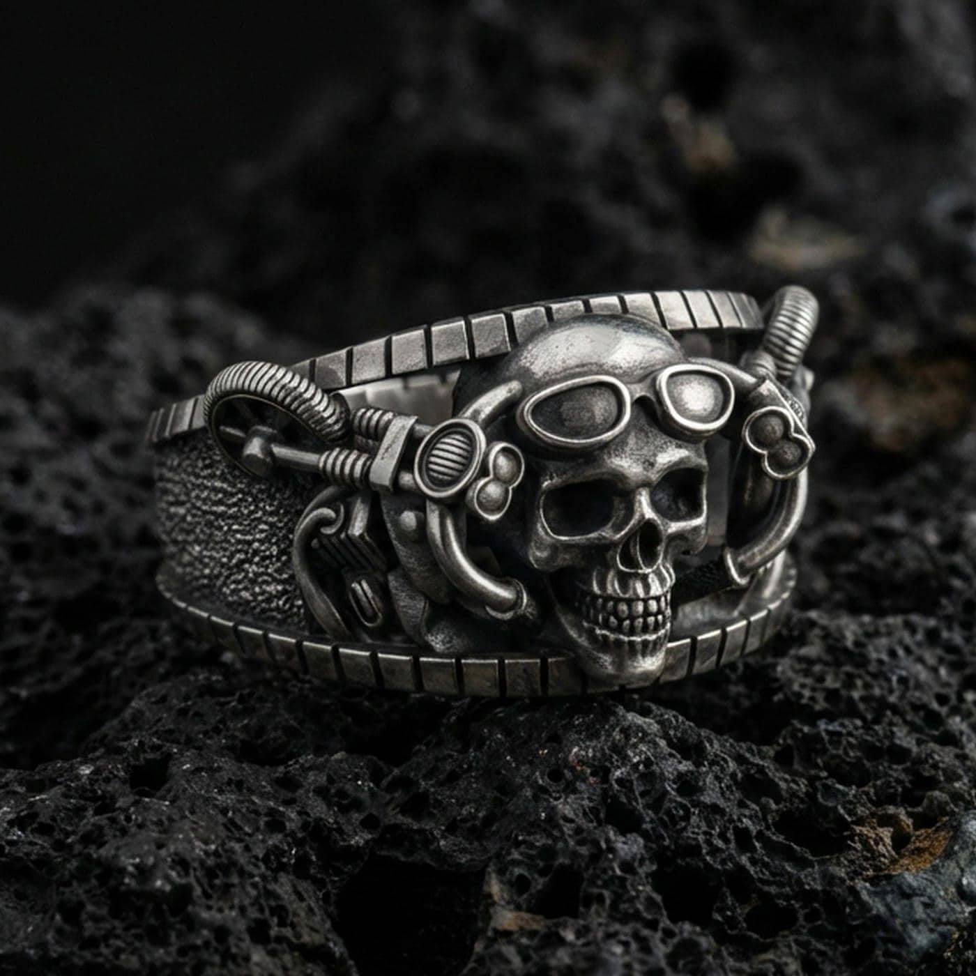 Oxidized sterling silver steampunk skull ring on dark volcanic rock, goggles and mechanical tubing detail, premium macro product shot.