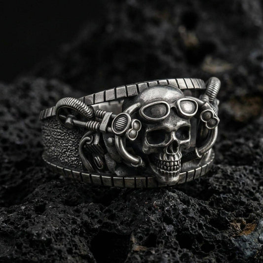Oxidized sterling silver steampunk skull ring on dark volcanic rock, goggles and mechanical tubing detail, premium macro product shot.