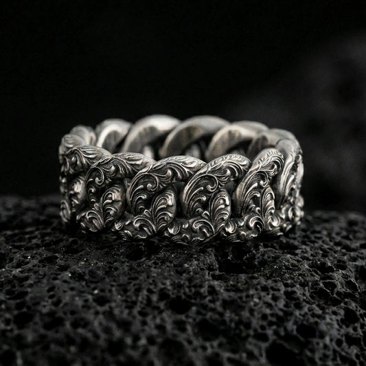 Oxidized sterling silver Cuban link chain ring on dark volcanic rock, engraved scrollwork on each link, premium macro product shot.