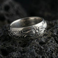 Load image into Gallery viewer, Oxidized sterling silver floral scroll band ring on dark volcanic rock, raised flower motif with flowing leaf engraving, premium macro product shot.

