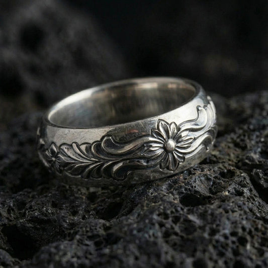 Oxidized sterling silver floral scroll band ring on dark volcanic rock, raised flower motif with flowing leaf engraving, premium macro product shot.