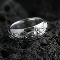 Load image into Gallery viewer, Polished silver floral engraved band ring on black volcanic stone, crisp flower relief and smooth swirling leaf pattern, centered close-up studio lighting.
