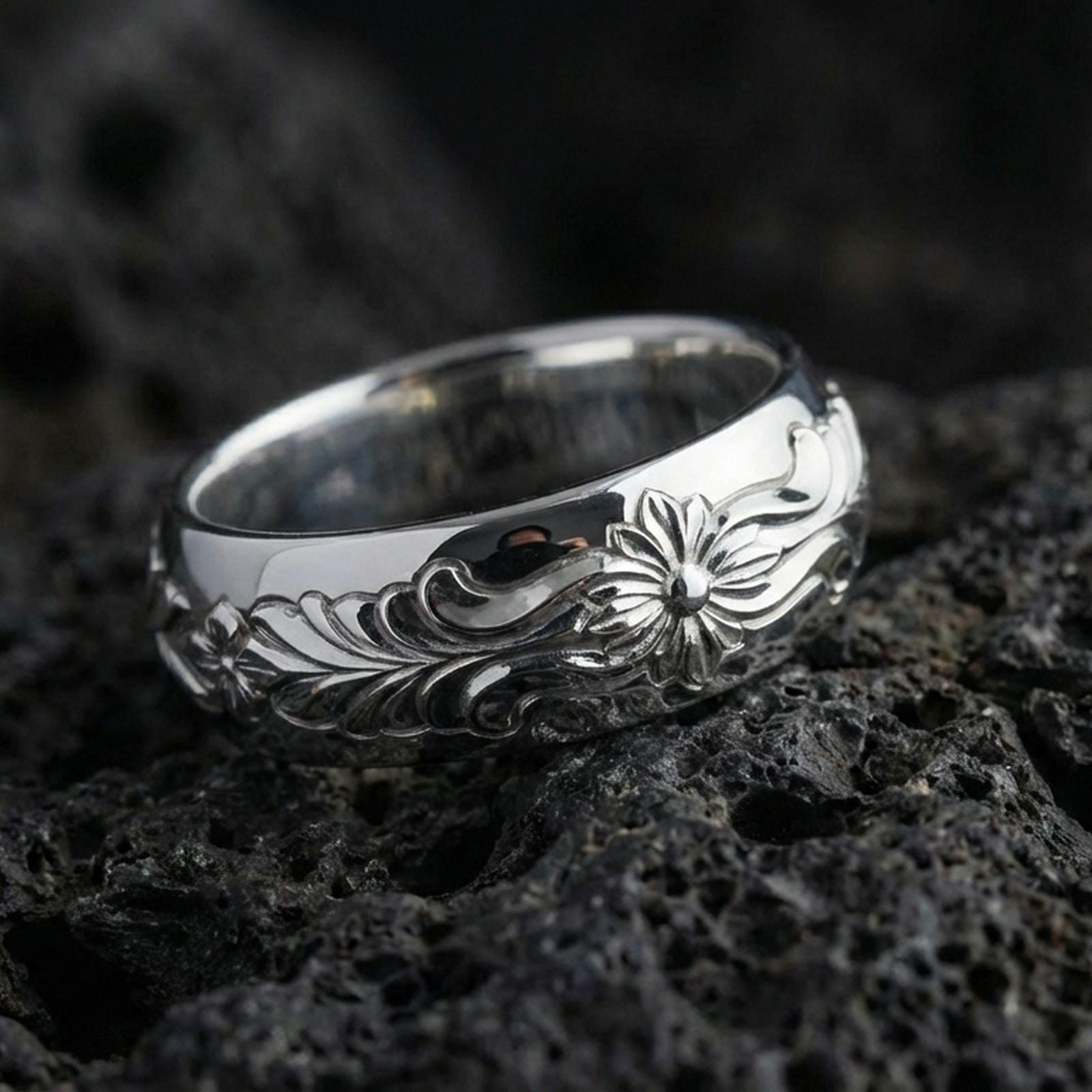 Polished silver floral engraved band ring on black volcanic stone, crisp flower relief and smooth swirling leaf pattern, centered close-up studio lighting.
