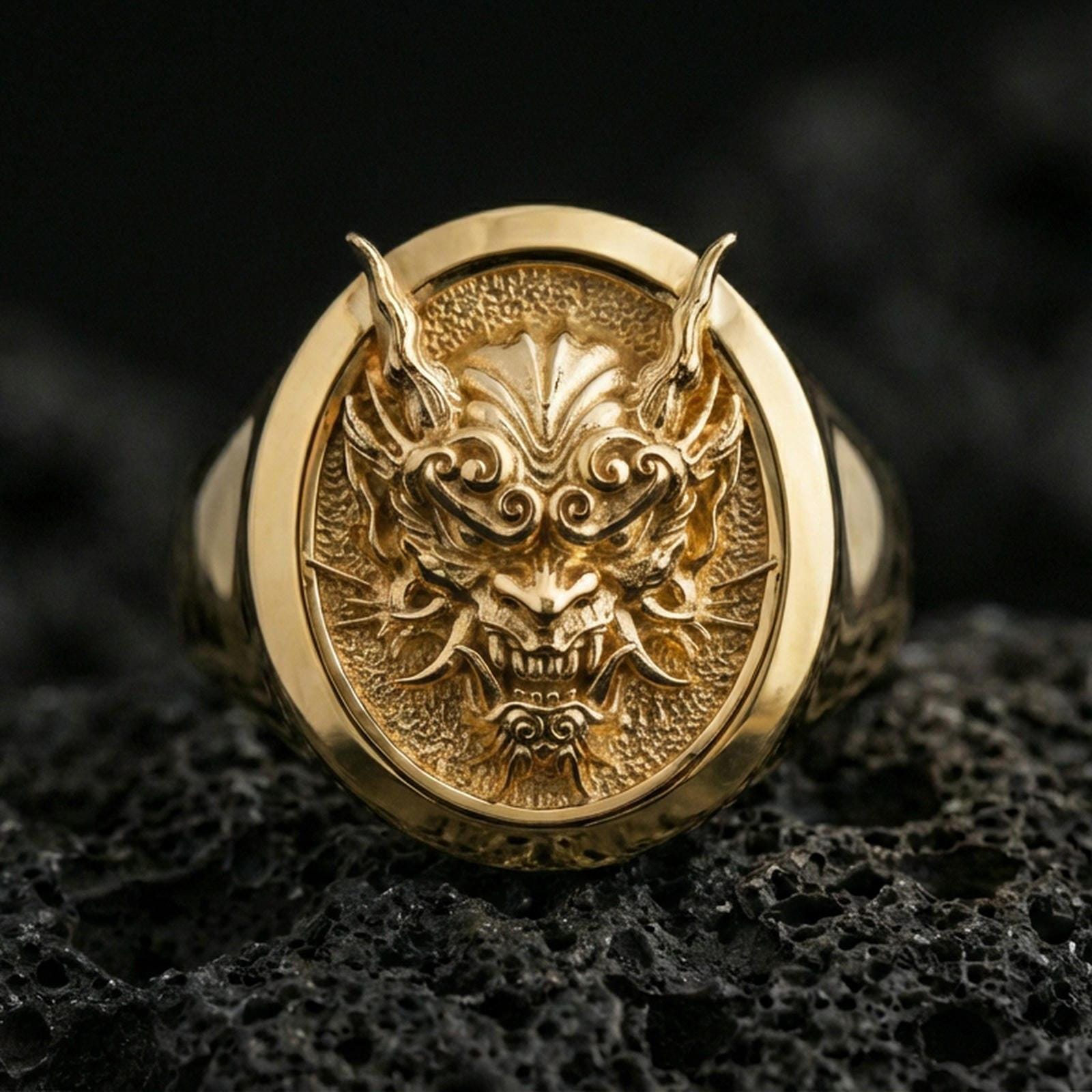 Gold plated oni mask ring on black volcanic rock, detailed view of the dramatic facial relief and sharp contours