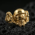 Load image into Gallery viewer, Gold plated biker skull ring on volcanic rock, detailed helmet, goggles, and rugged skull relief, luxury jewelry product photography.
