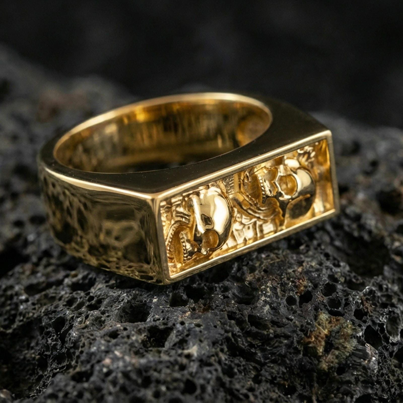 Sterling silver minimalist signet ring with a gold plated finish, rectangular open frame with tiny skull detailing and industrial texture, macro on volcanic rock