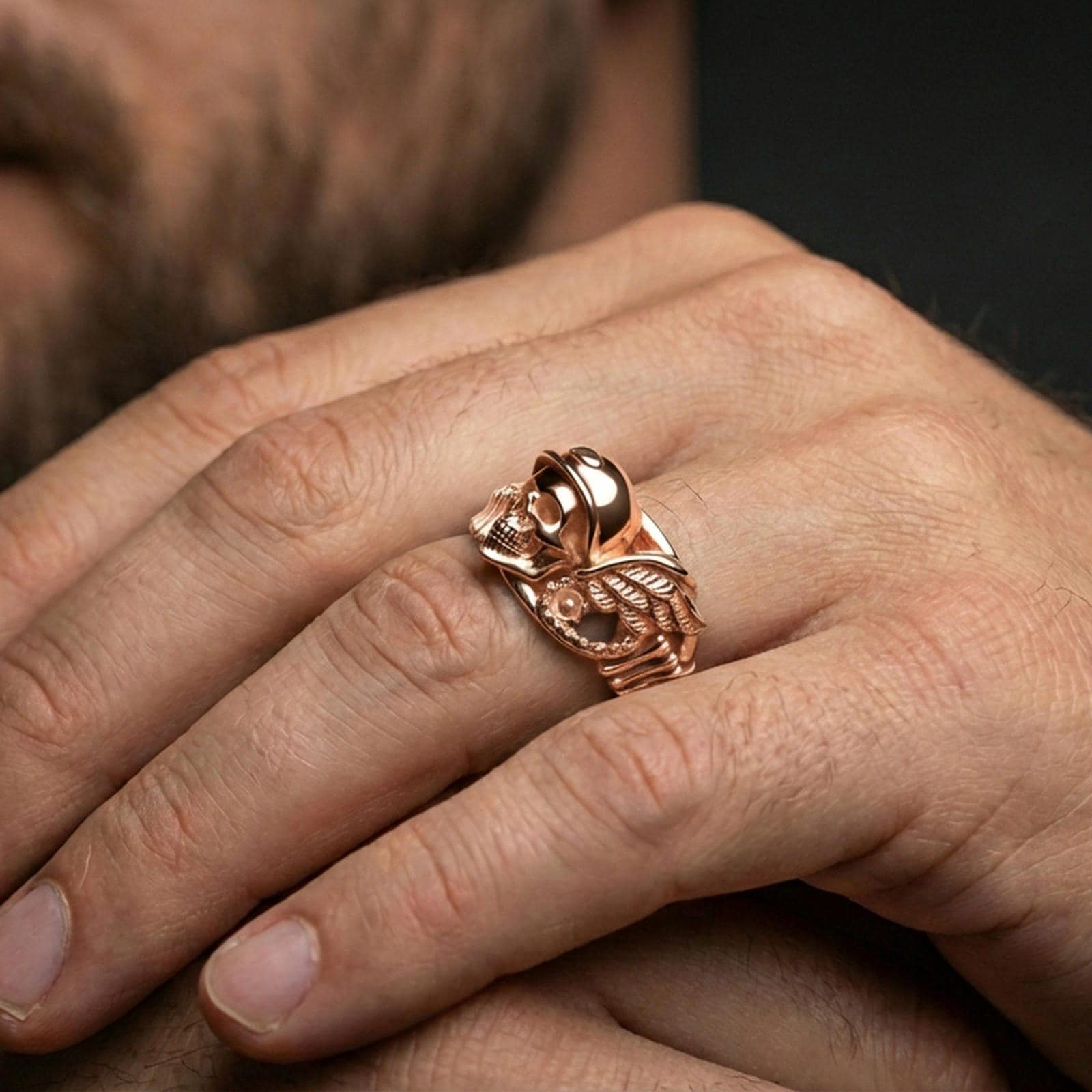Rose gold plated gothic biker skull ring worn on finger, sculpted helmeted skull relief with wing detail, elegant lifestyle wear shot.