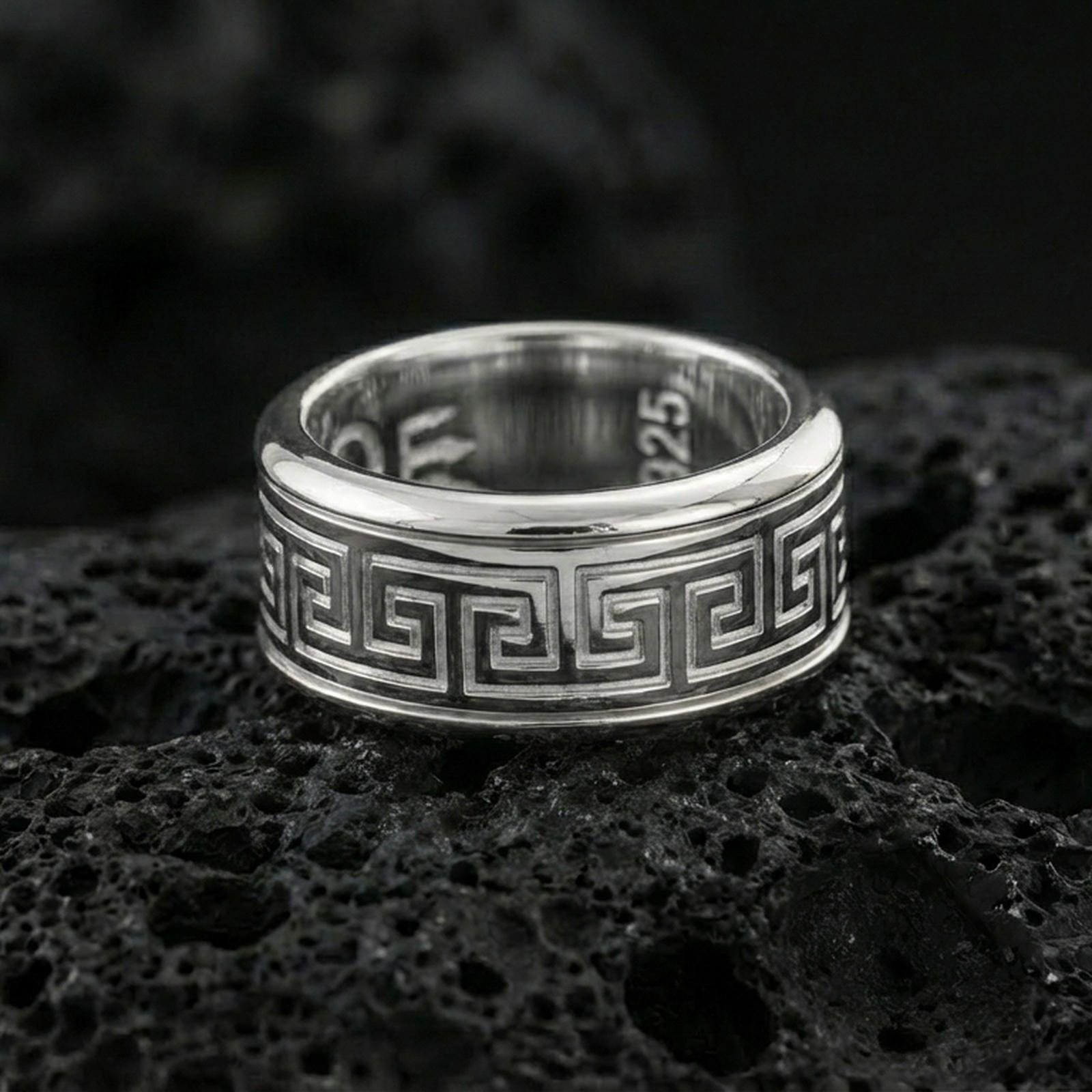 Polished silver Greek key wedding band on black volcanic stone, crisp geometric meander border detail, centered close-up studio lighting.