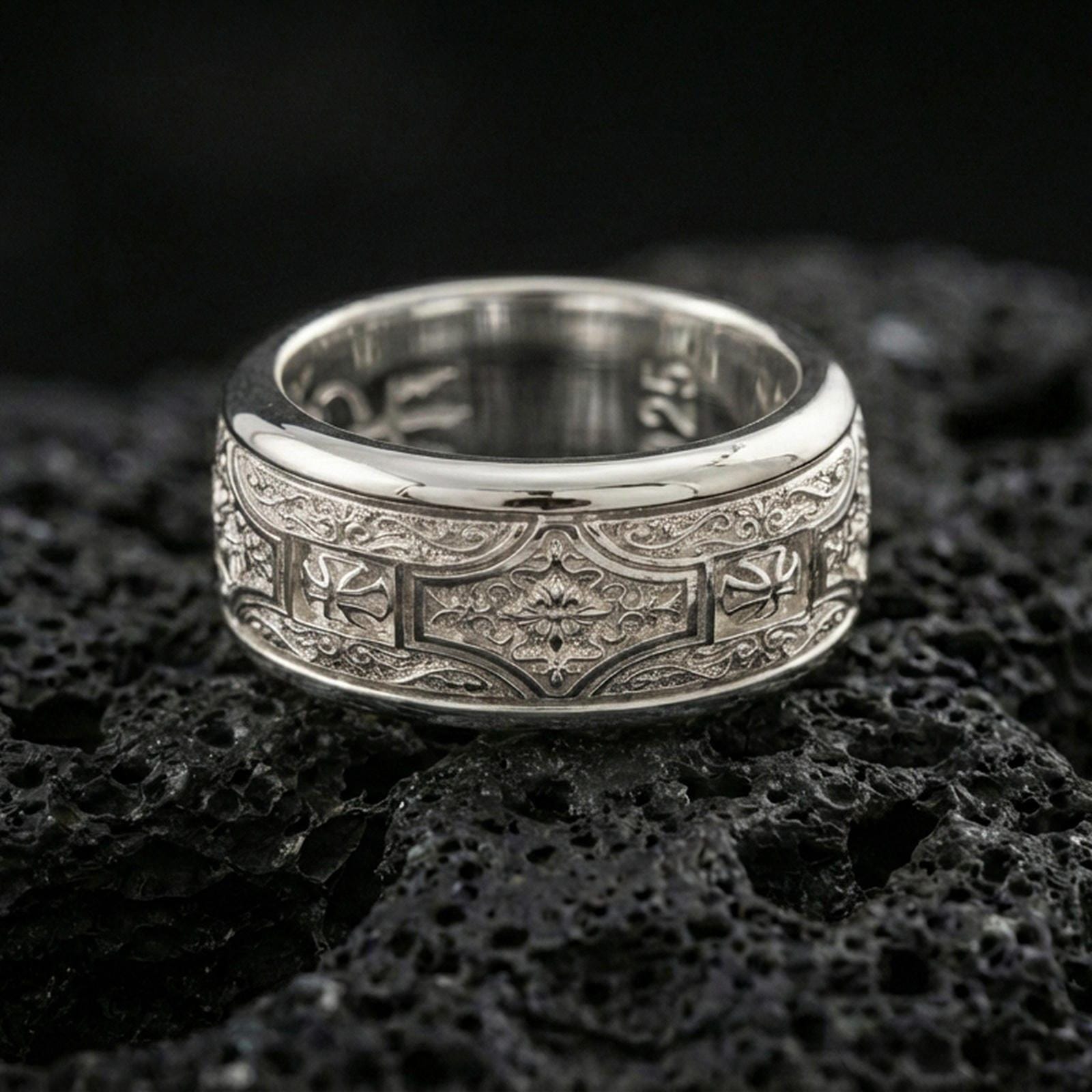 Polished silver engraved ornament band ring on black volcanic stone, crisp floral medallion panel with detailed scrollwork borders, centered close-up studio lighting.
