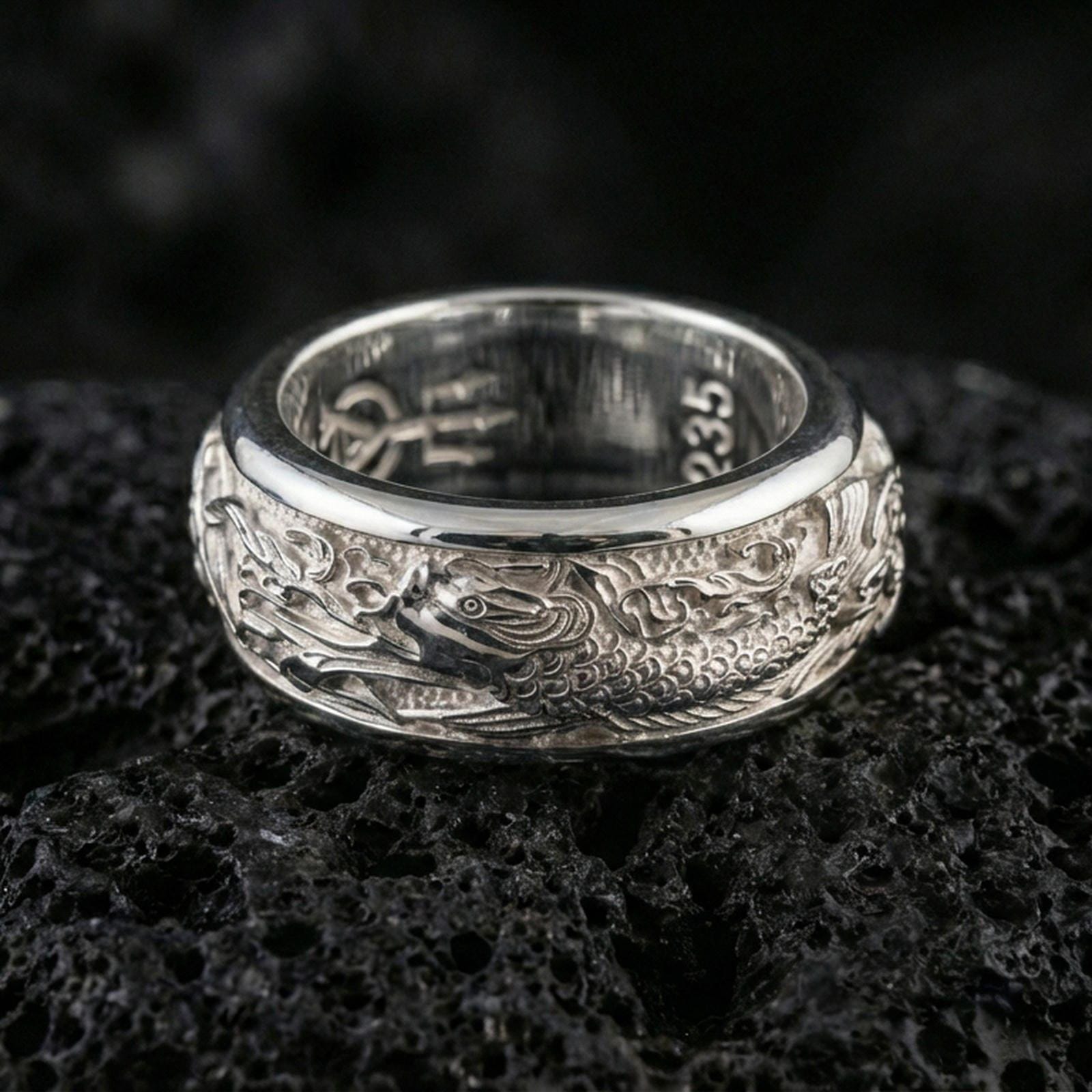 Polished silver koi fish fidget spinner ring on black volcanic stone, crisp koi scale relief with flowing wave pattern, centered close-up studio lighting.