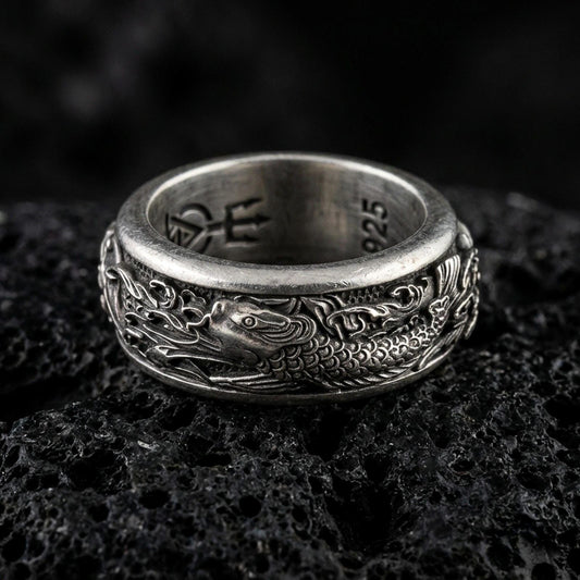 Oxidized sterling silver koi fish spinner band ring on dark volcanic rock, detailed koi and wave engraving on rotating center, premium macro product shot.