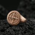 Load image into Gallery viewer, Sterling silver compass signet ring with a rose gold plated finish, bold round face featuring compass rose star and cardinal direction engraving, close-up on volcanic rock

