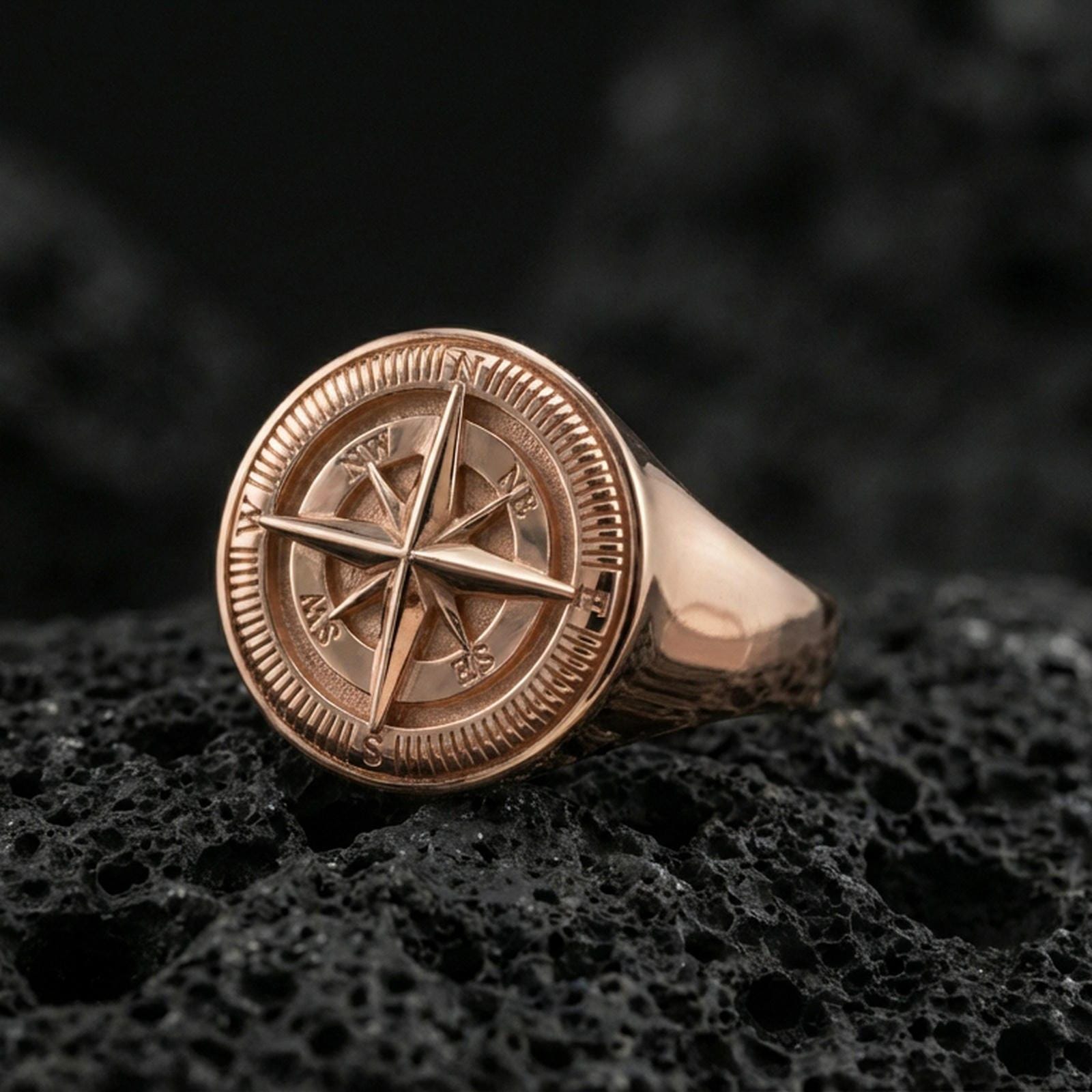 Sterling silver compass signet ring with a rose gold plated finish, bold round face featuring compass rose star and cardinal direction engraving, close-up on volcanic rock