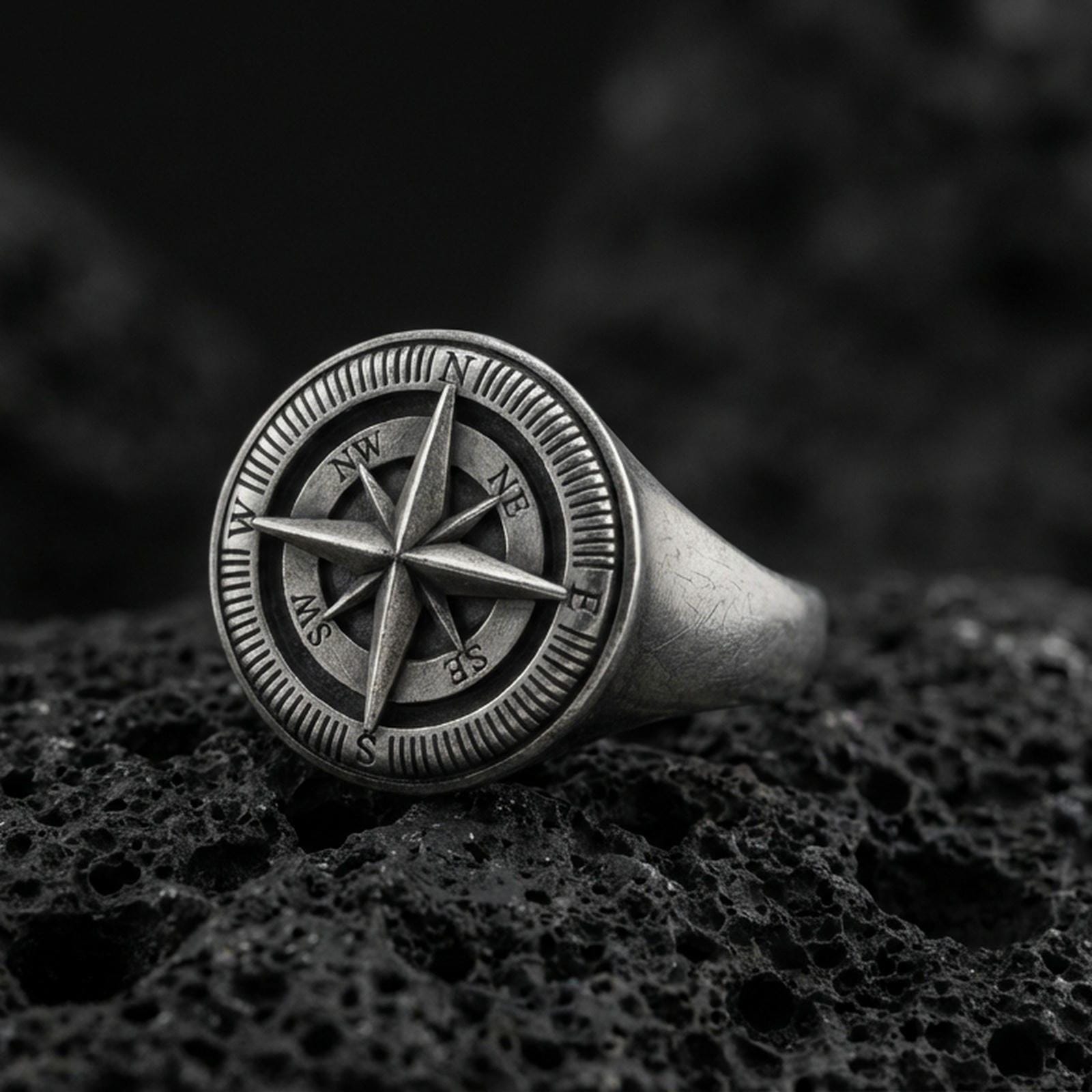 Sterling silver compass rose signet ring with an oxidized finish, engraved cardinal directions and nautical star design, macro on black volcanic rock