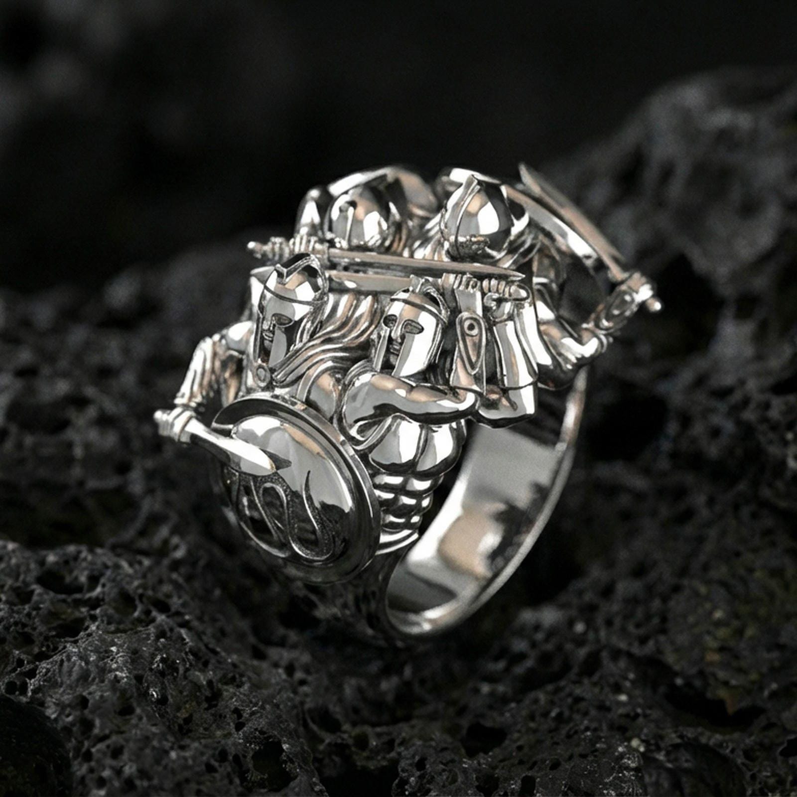 Sterling silver Greek warrior statement ring in a polished silver finish, high-relief Spartan soldiers design with helmets, shield, and raised weapons, close-up on volcanic stone