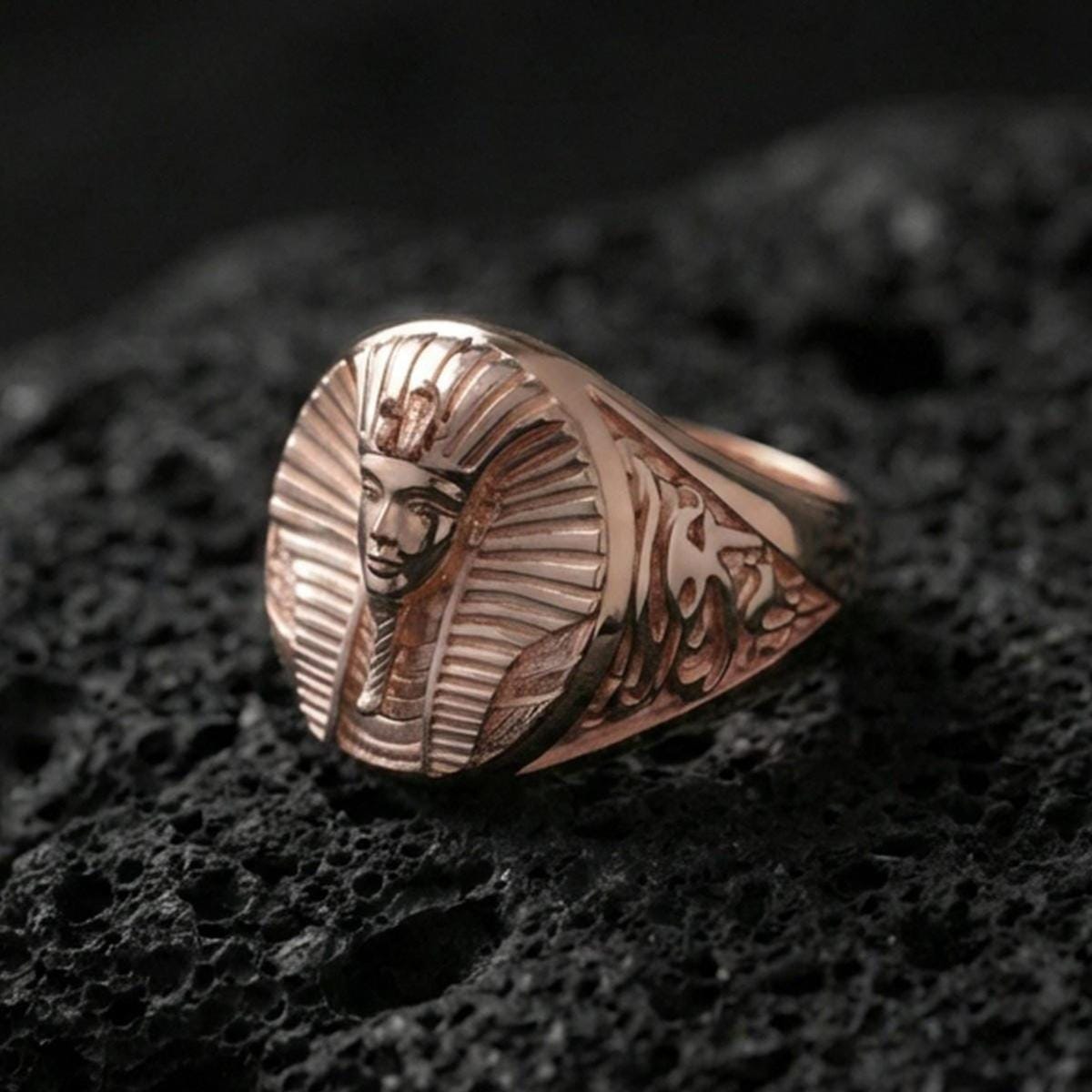 Sterling silver ancient Egypt signet ring with a rose gold plated finish, oval pharaoh portrait face and carved shoulder accents, close-up on black volcanic rock