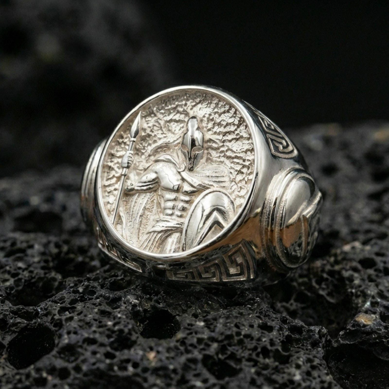 Sterling silver Spartan warrior ring in a polished silver finish, bold round signet design with detailed Greek hoplite relief and carved side accents, close-up on volcanic stone