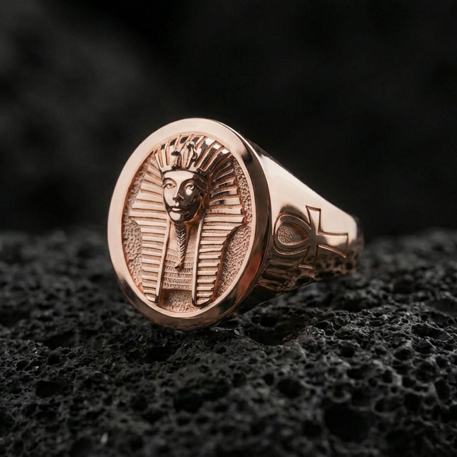 Sterling silver Egyptian king signet ring with a rose gold plated finish, bold oval statement face with pharaoh bust engraving and symbolic ankh detail, close-up on black volcanic rock
