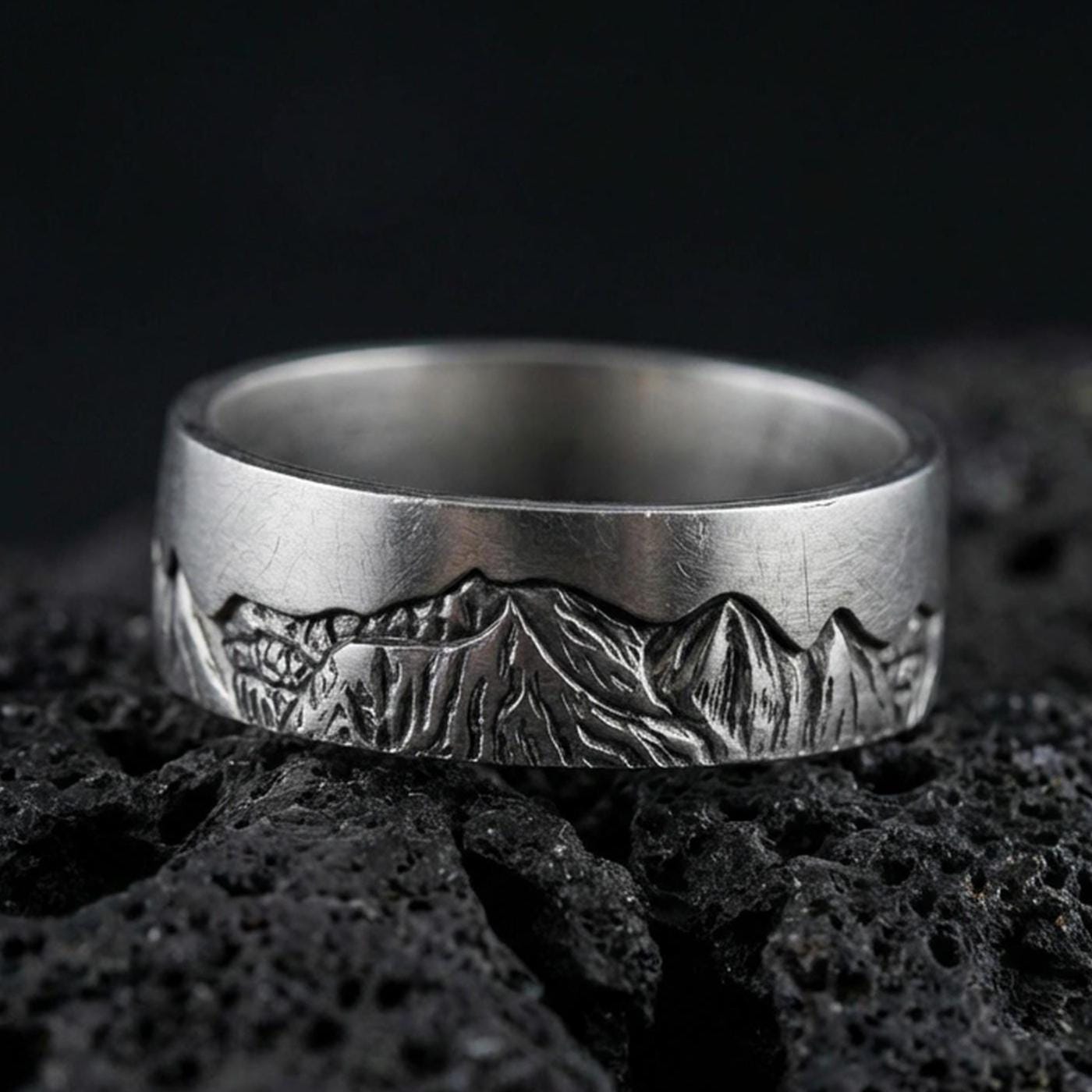 Oxidized sterling silver Grand Teton mountain band ring on dark volcanic rock, detailed alpine skyline engraving, premium macro product shot.